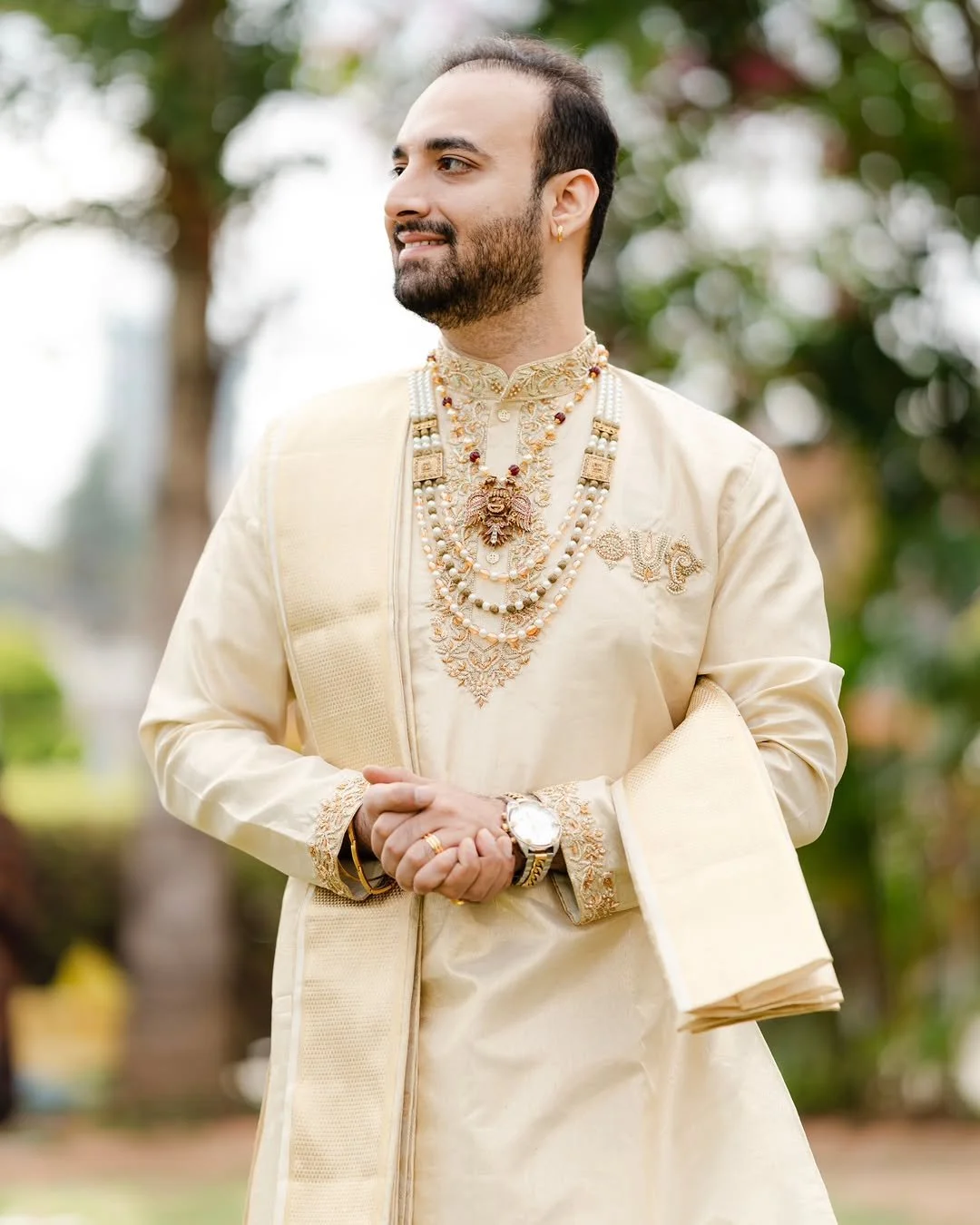 Man wearing traditional Indian cream-colored embroidered clothing and multiple pearl necklaces, standing outdoors with trees in the background.