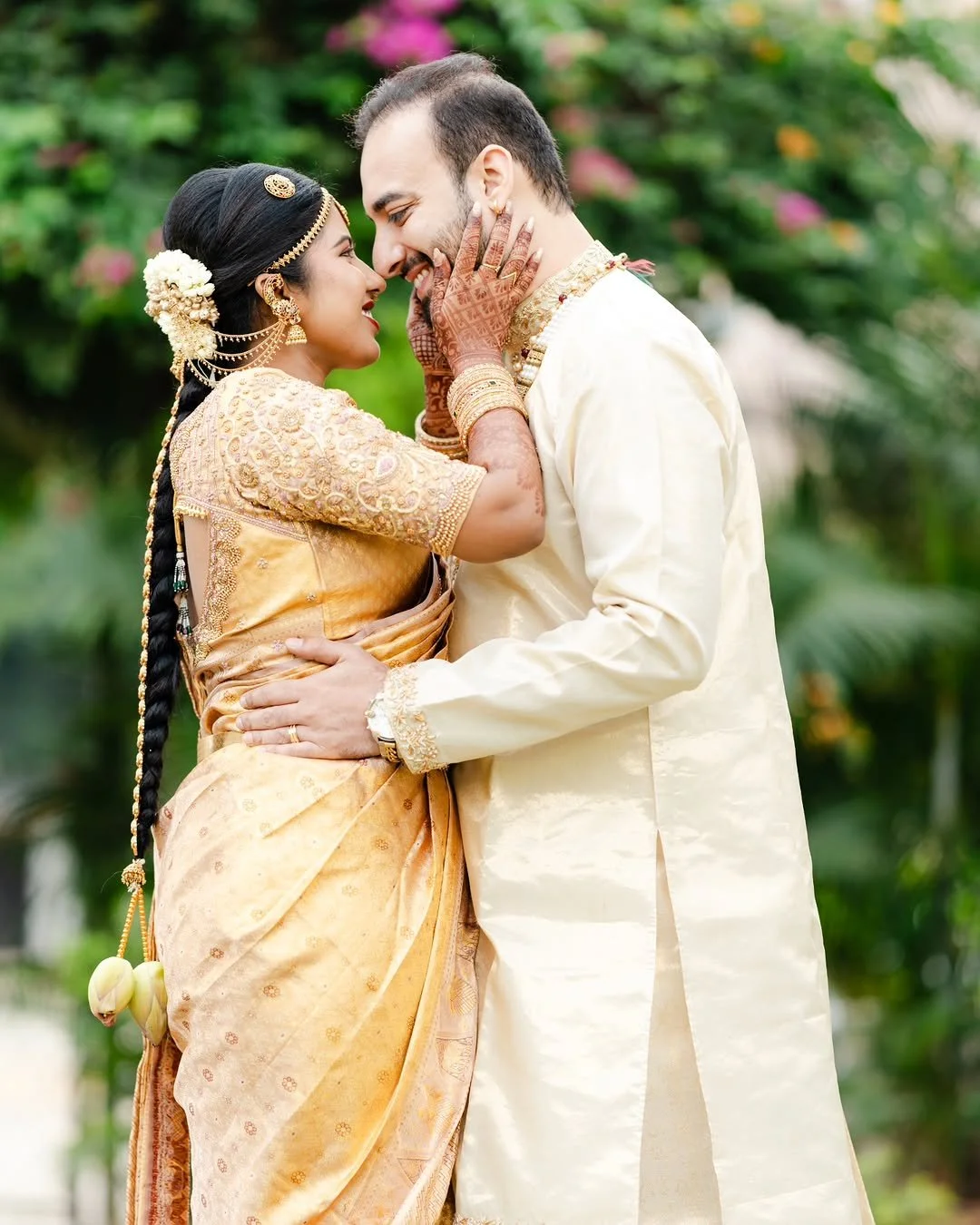 A couple dressed in traditional Indian wedding attire, embracing outdoors with greenery and colorful flowers in the background.