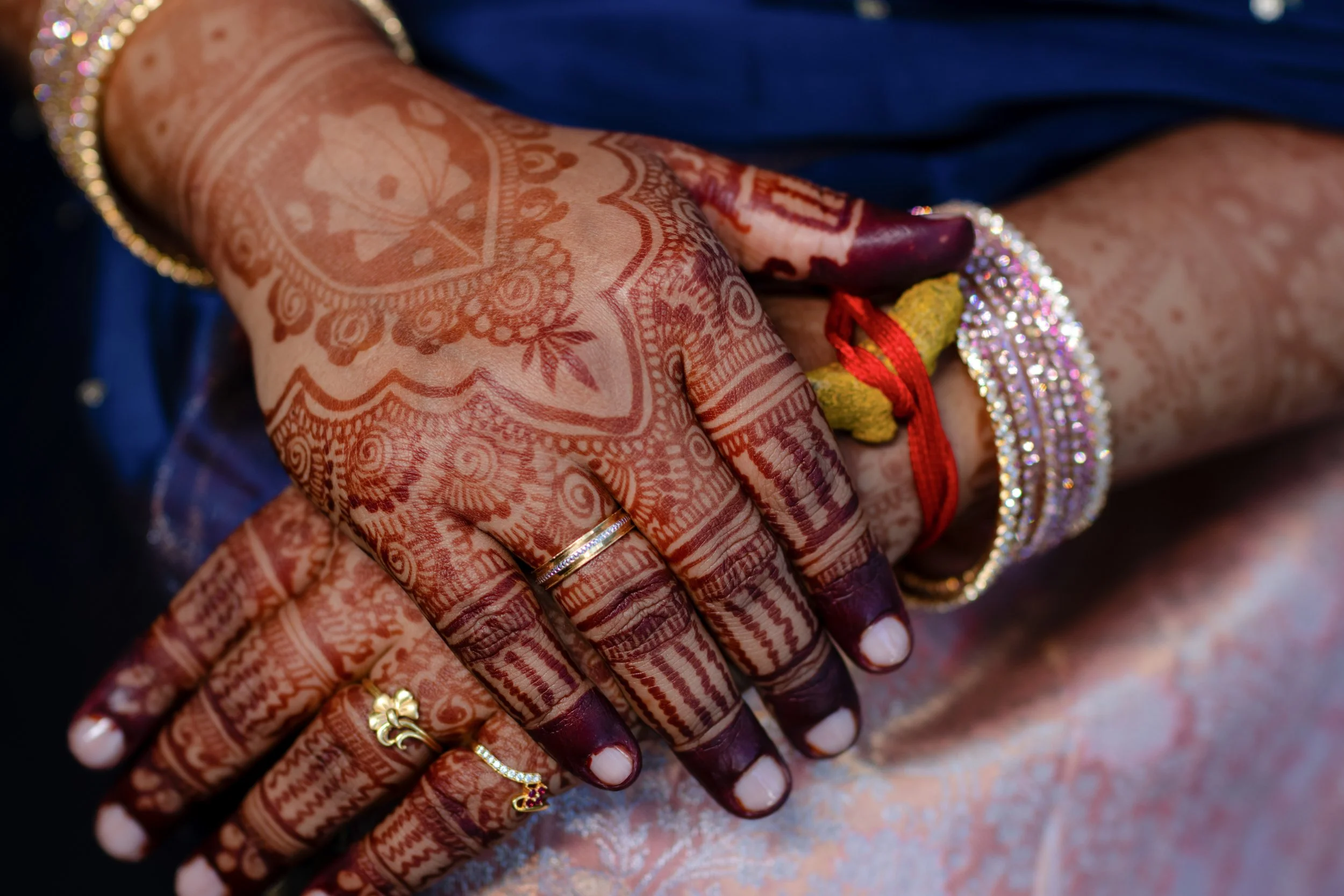 Close-up of a woman's hand adorned with intricate henna designs, multiple gold rings, and colorful bangles, with a dark blue sleeve in the background.