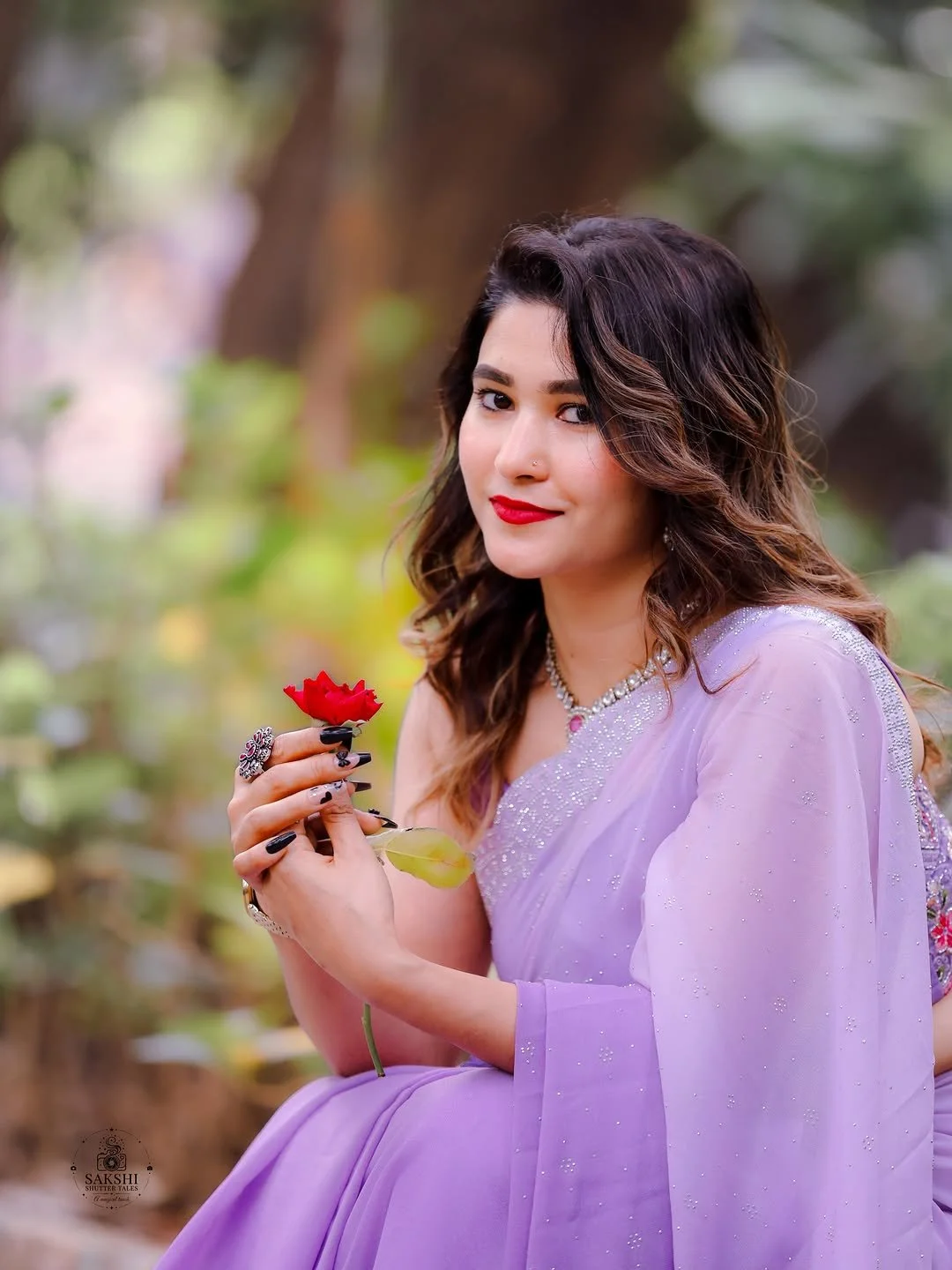 A woman with wavy brown hair and red lipstick, wearing a lavender saree with embellishments, sitting outdoors and holding a red flower.