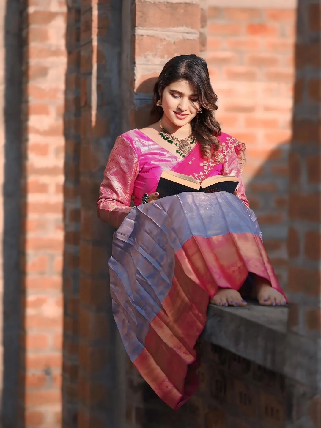 A woman wearing a pink and purple traditional Indian saree, sitting on a ledge, reading a book, with a brick wall background.