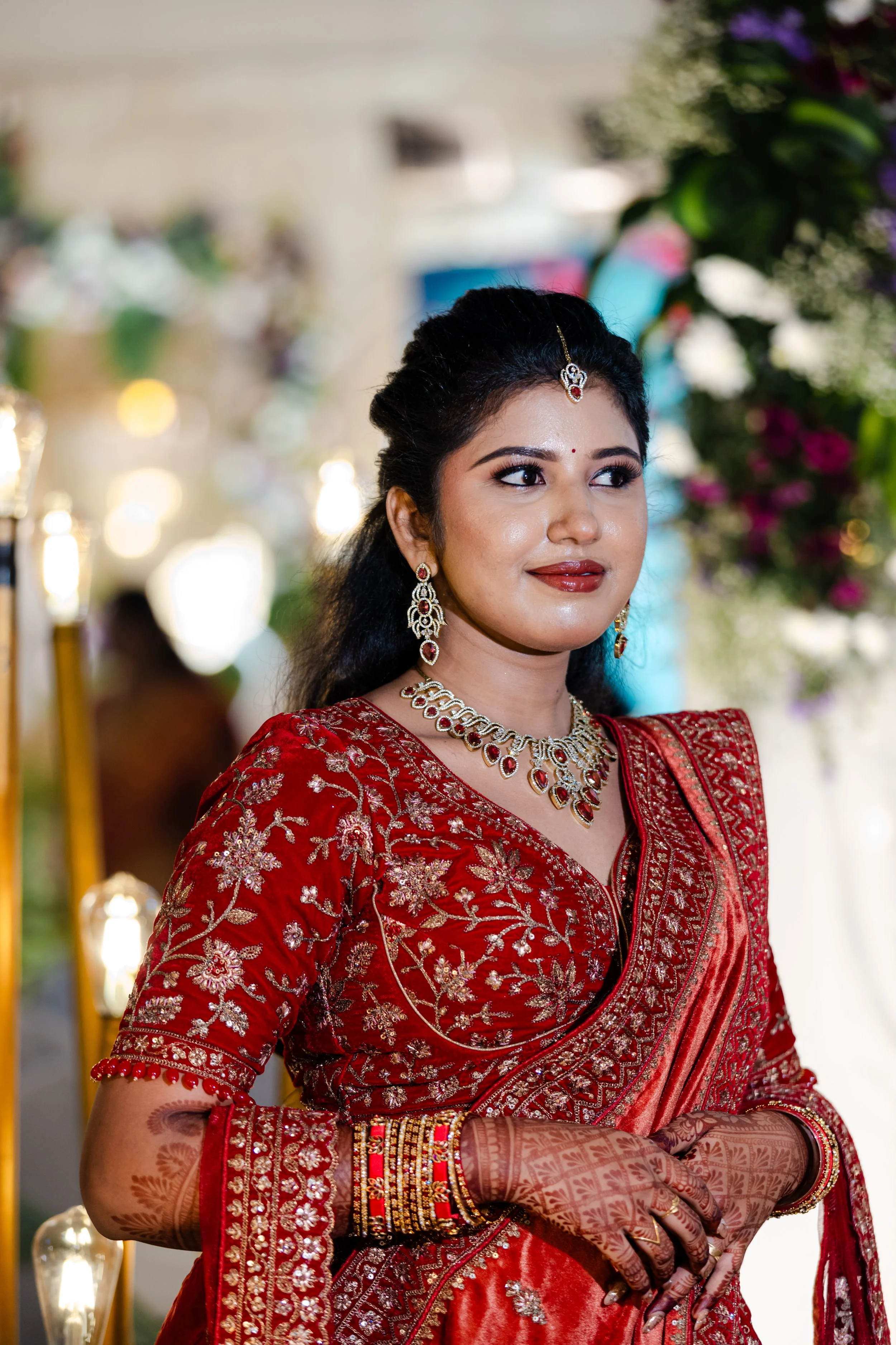A woman in traditional Indian attire, wearing a red embroidered saree, ornate jewelry including earrings, a necklace, and a maang tikka, with henna designs on her hands and smiling while standing in a decorated setting with flowers and lights.