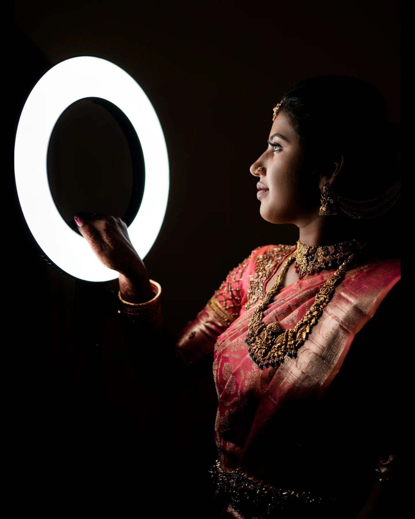A woman dressed in traditional Indian attire, jewelry, and makeup holding a ring light, with her face in profile.
