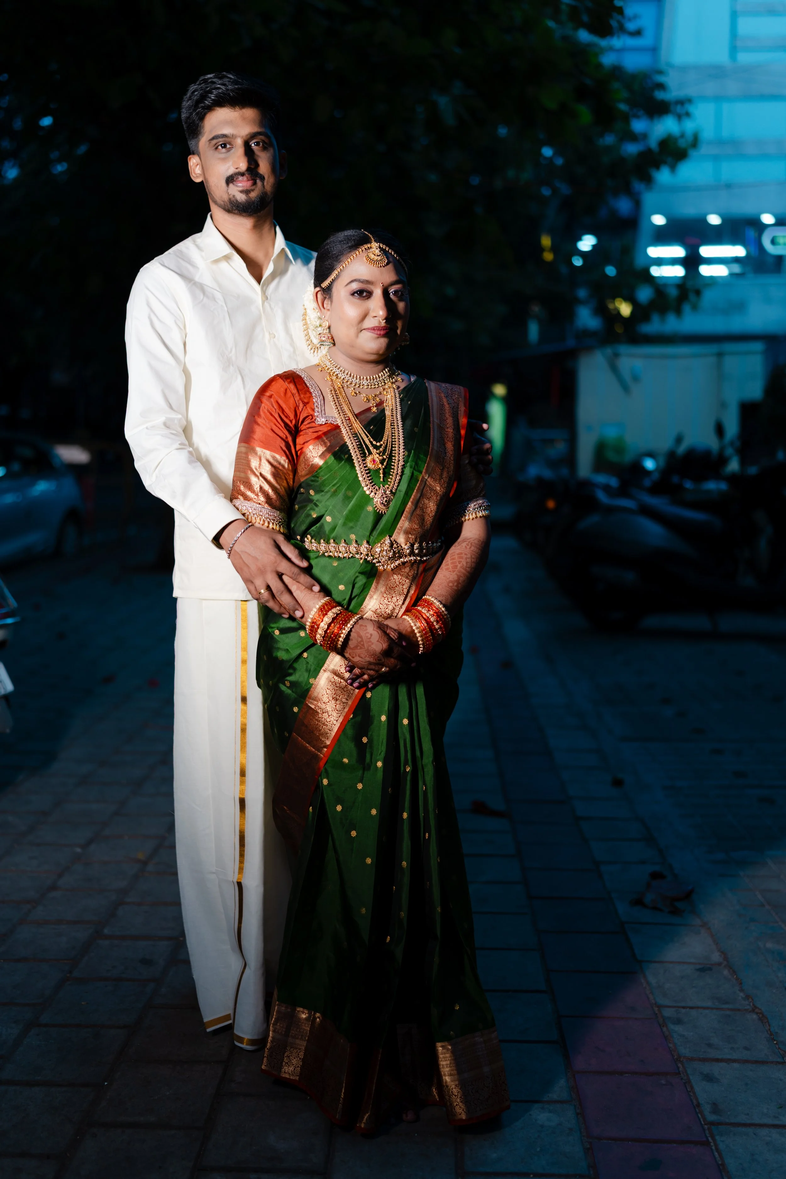A man and woman dressed in traditional Indian wedding attire standing outdoors at night. The man is wearing a white kurta with gold accents, and the woman is wearing a green saree with gold jewelry, including a necklace, earrings, and headpiece.