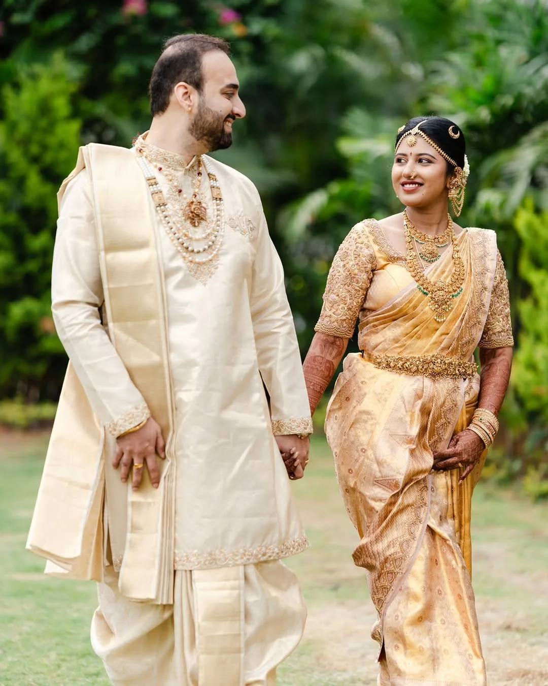A couple dressed in traditional Indian wedding attire walking outdoors, holding hands and smiling at each other.