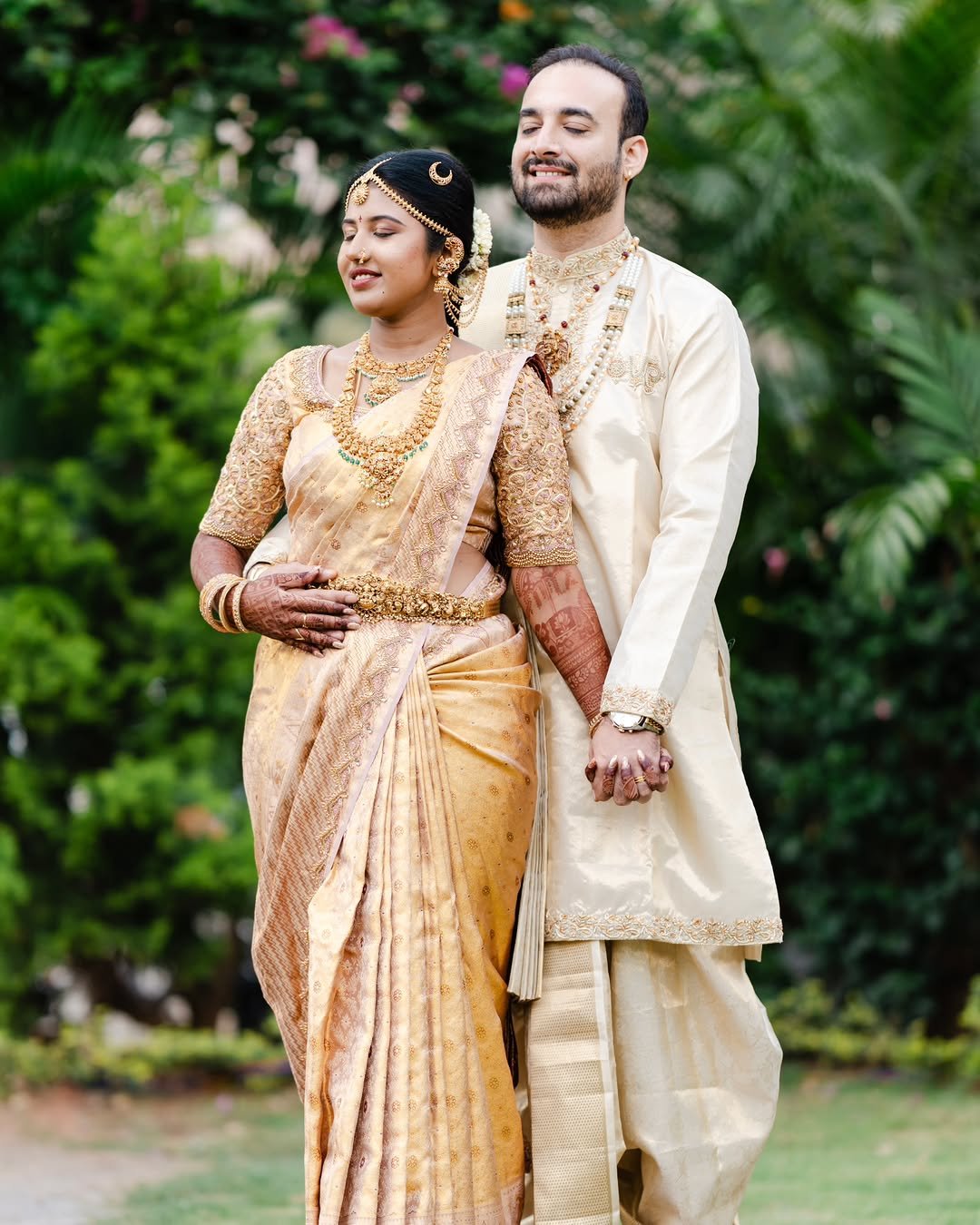 A couple dressed in traditional Indian wedding attire, holding hands outdoors in a garden setting with greenery and pink flowers in the background.