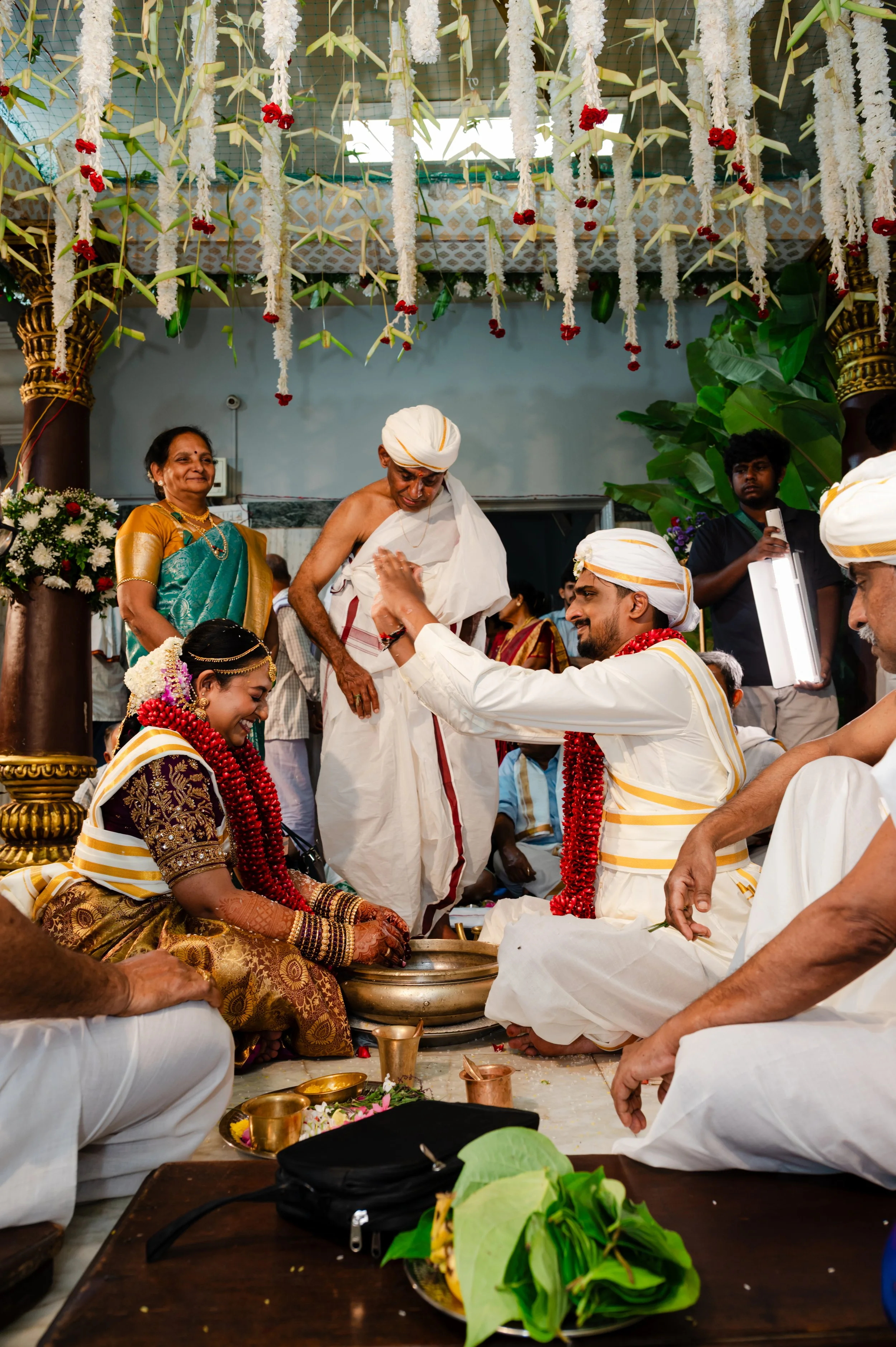 A traditional Indian wedding ceremony with the bride and groom seated on the floor, surrounded by family members. The bride is dressed in a richly embroidered saree with gold jewelry, and the groom in white traditional attire with a turban. They are 
