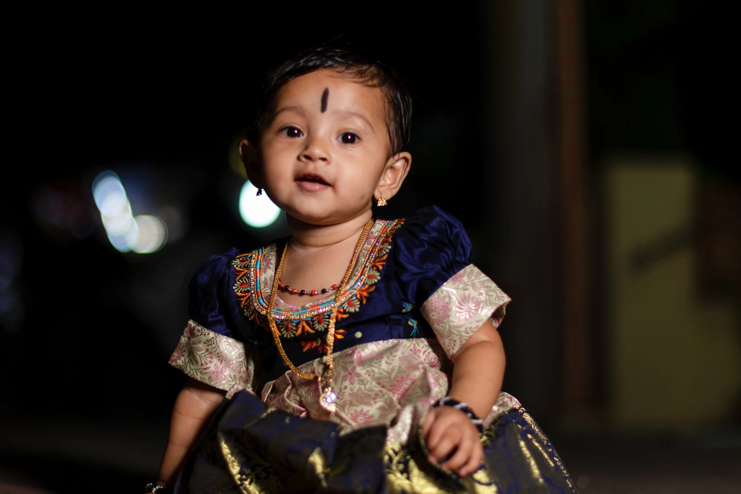 Young girl wearing traditional Indian dress and jewelry, with a black bindi on her forehead, during nighttime with blurred lights in the background.