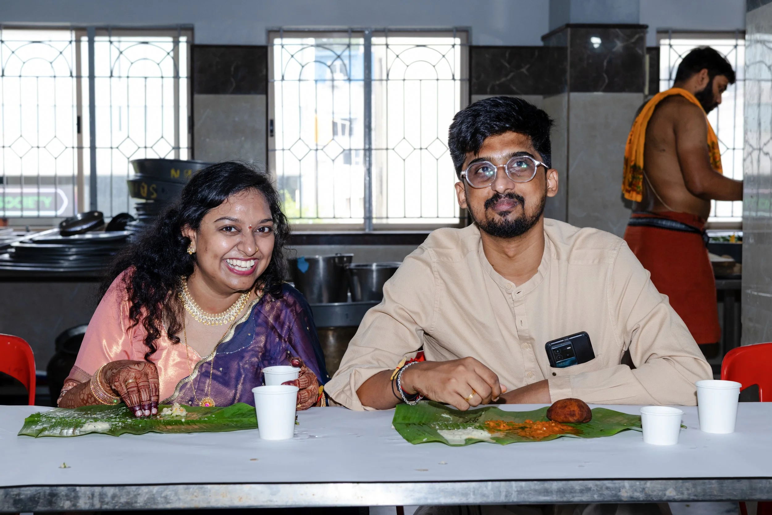 A couple of people sitting at a table during a meal, with the woman wearing traditional Indian attire and jewelry, and the man wearing glasses and a beige shirt. There are food items and cups on the table, with a man standing in the background prepar