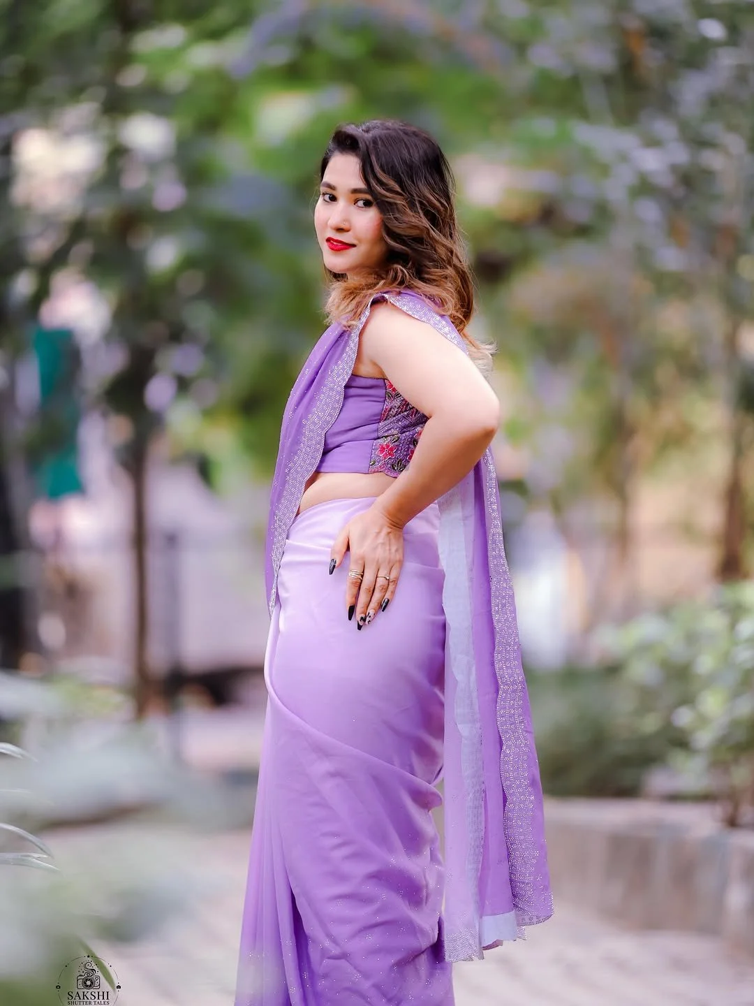 A woman in a purple saree standing outdoors with trees in the background, looking at the camera with one hand on her hip.