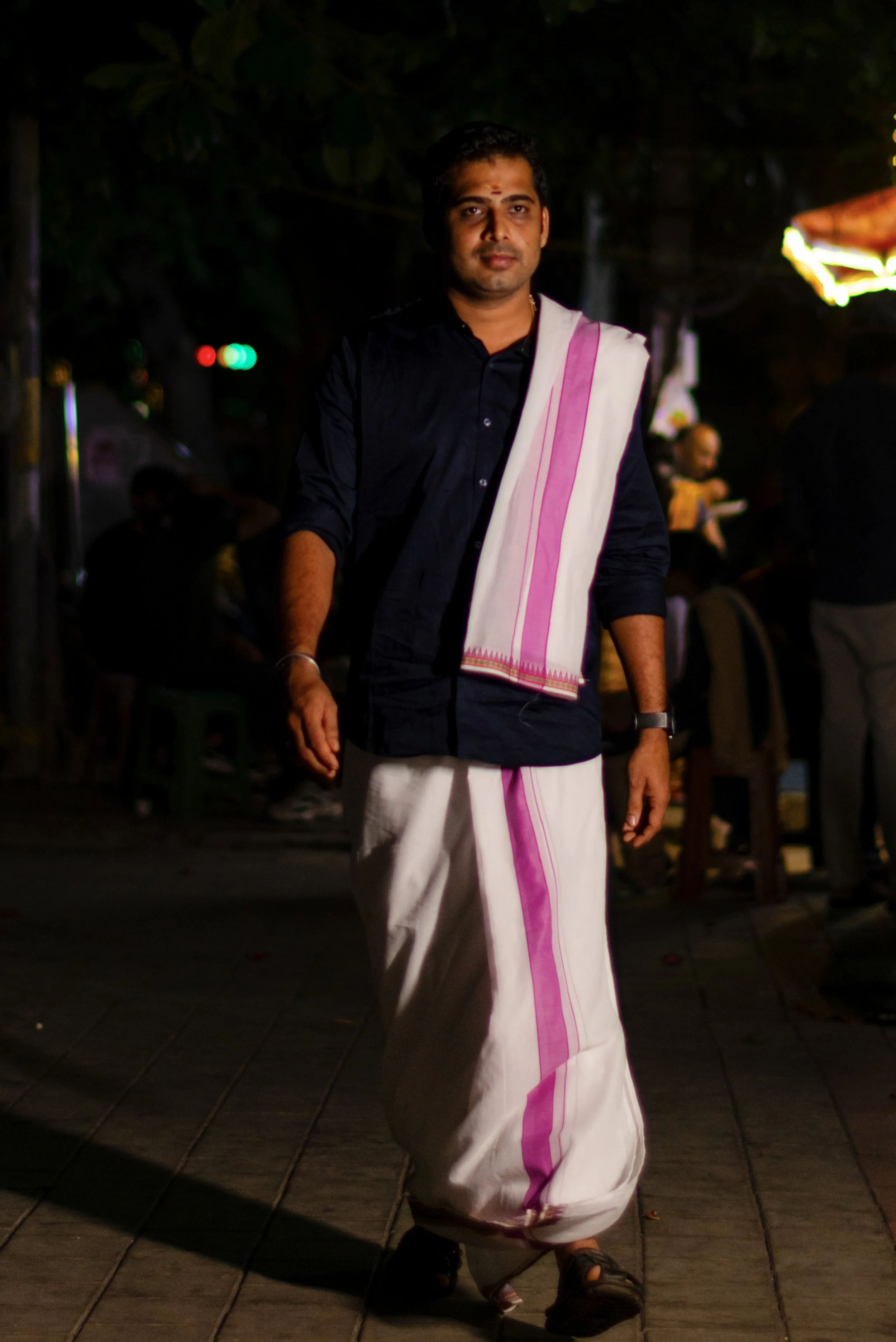A man wearing traditional Indian attire, including a black shirt, white dhoti with pink borders, and sandals, walking at night in a dimly lit outdoor setting with other people visible in the background.