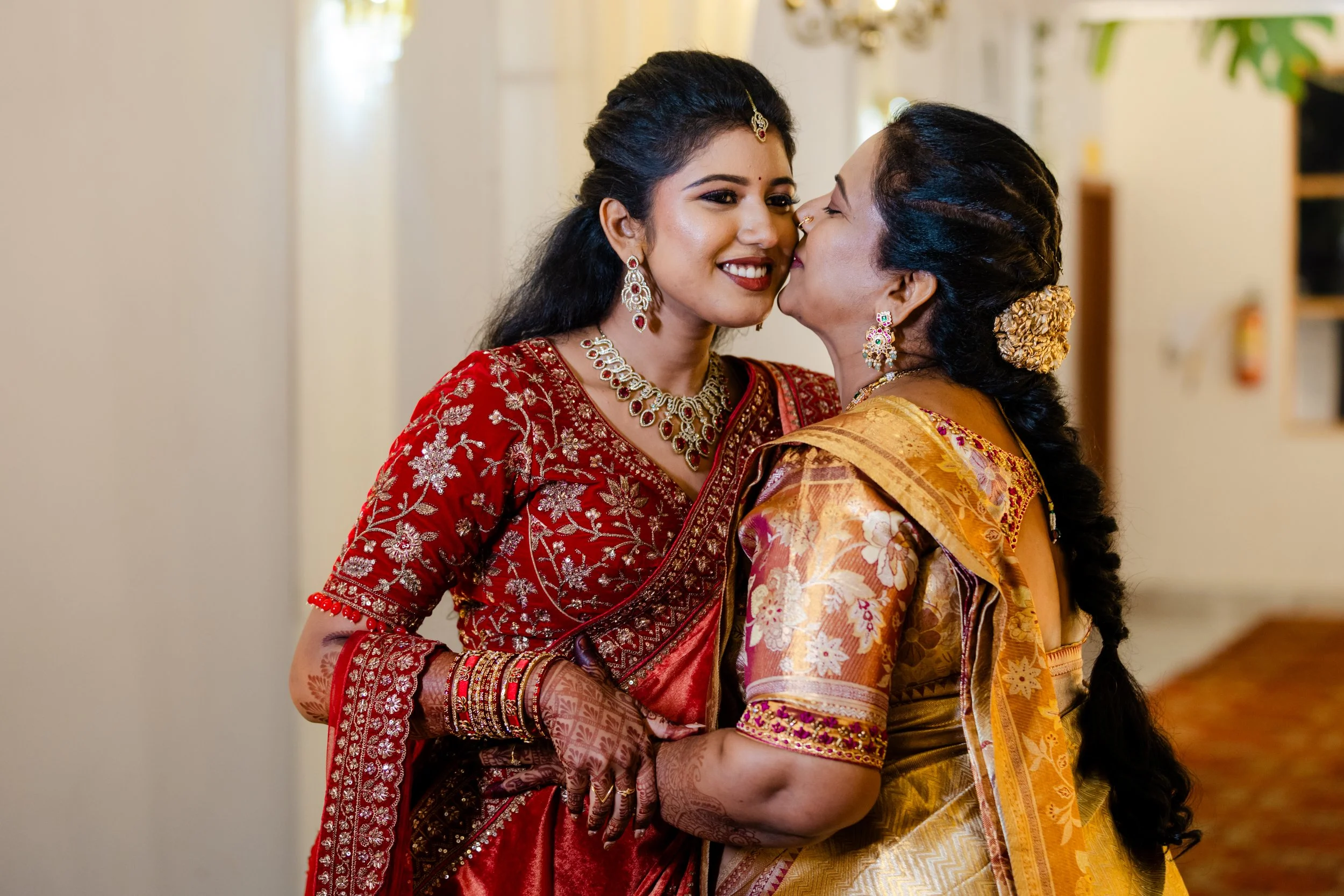 Two women dressed in traditional Indian attire sharing a close, affectionate moment, with one smiling and the other leaning in.