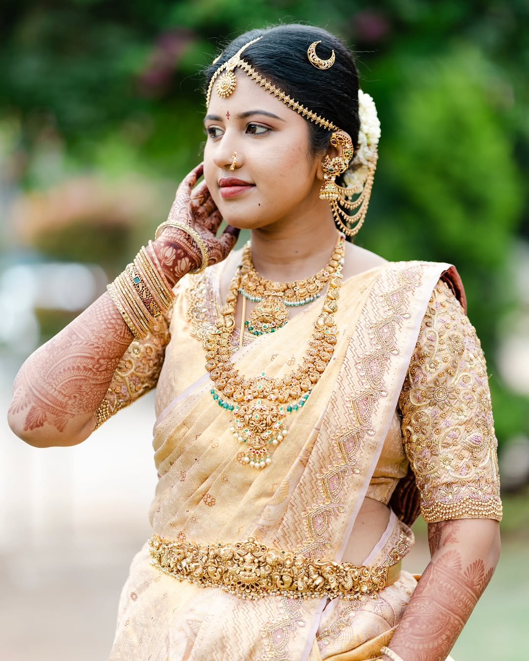 A woman dressed in traditional Indian wedding attire and jewelry, including a saree, necklaces, earrings, bangles, and a headpiece, standing outdoors with greenery in the background.