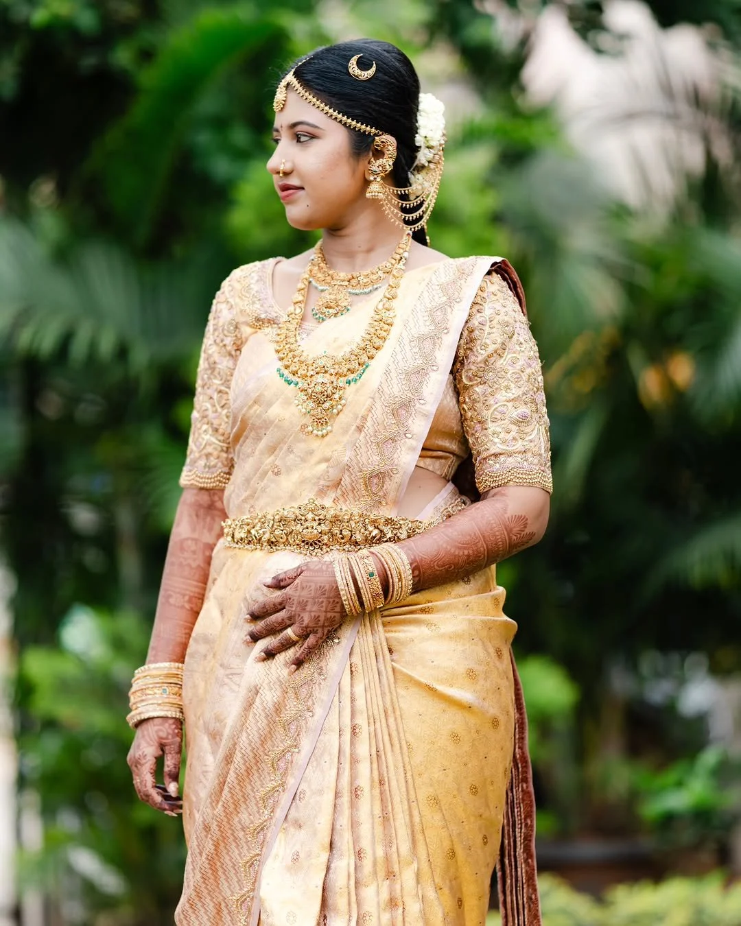 A woman dressed in traditional Indian attire wearing gold jewelry, bangles, and henna on her hands, standing outdoors with green foliage in the background.