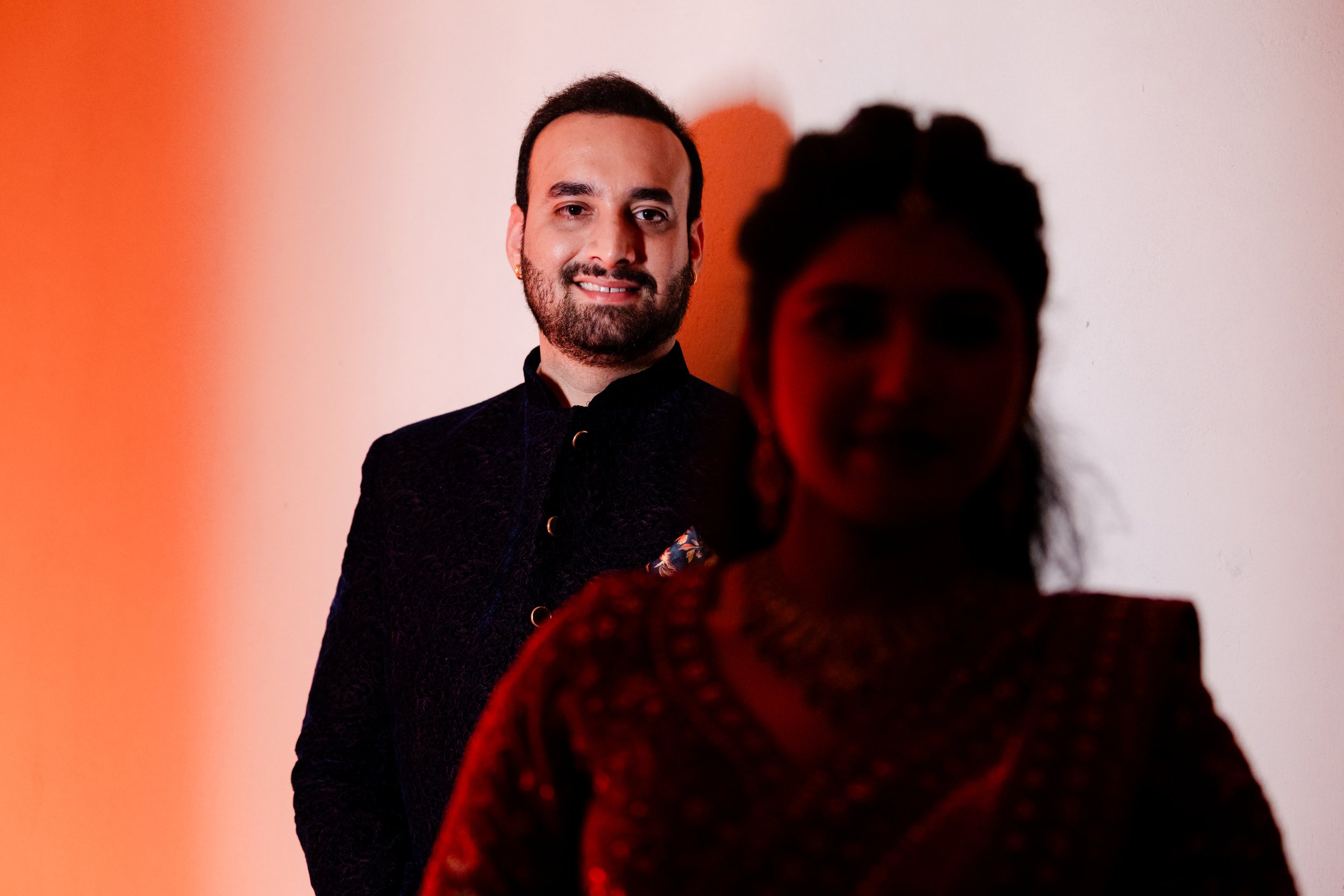 A man with dark hair, a beard, and earrings smiling, stands against a plain wall illuminated with warm light. In the foreground, a woman with dark, curly hair and earrings, dressed in red and black, is out of focus.
