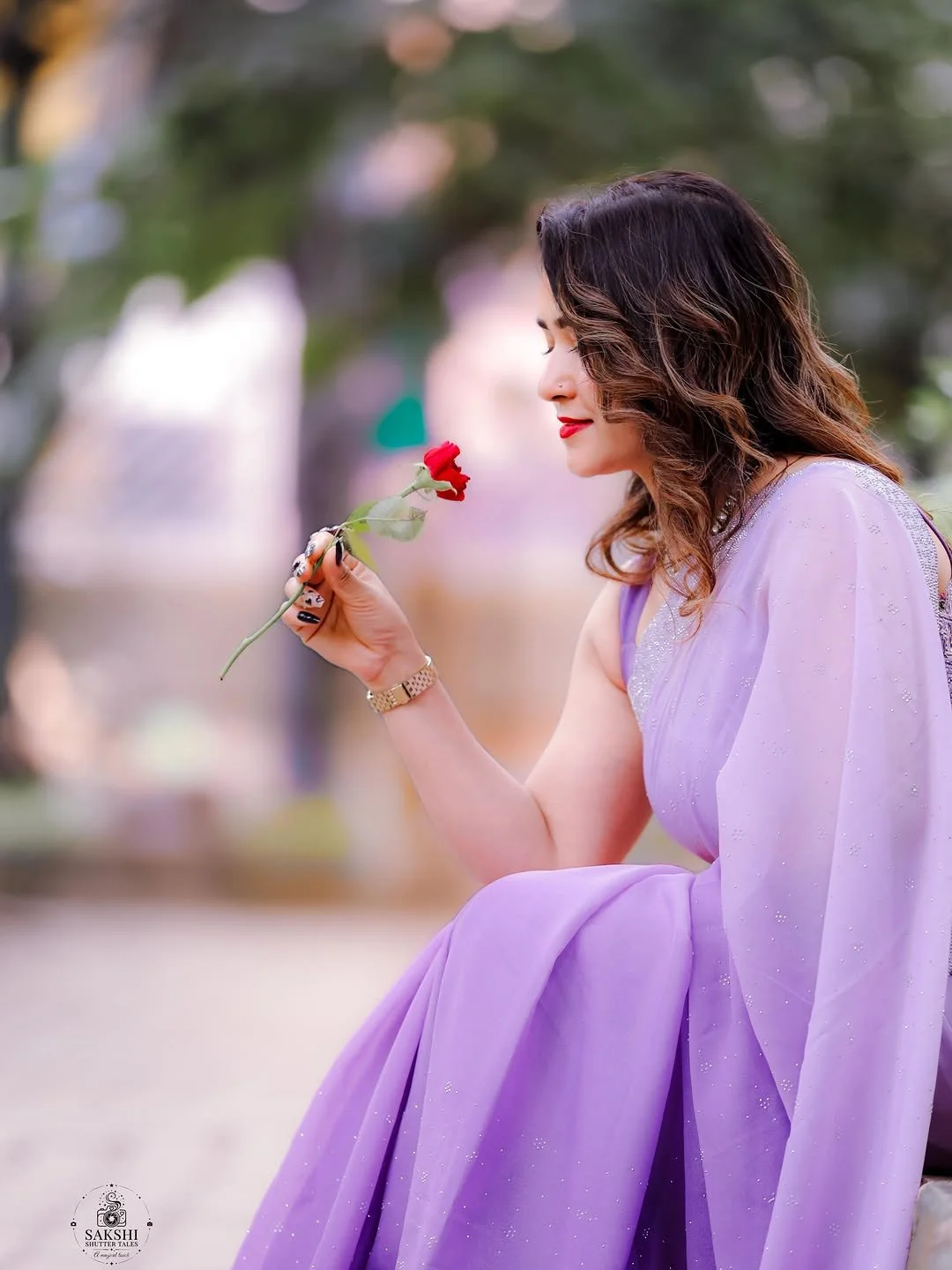 Woman with wavy brown hair and red lipstick holding a red carnation flower while sitting outdoors in a lavender saree.