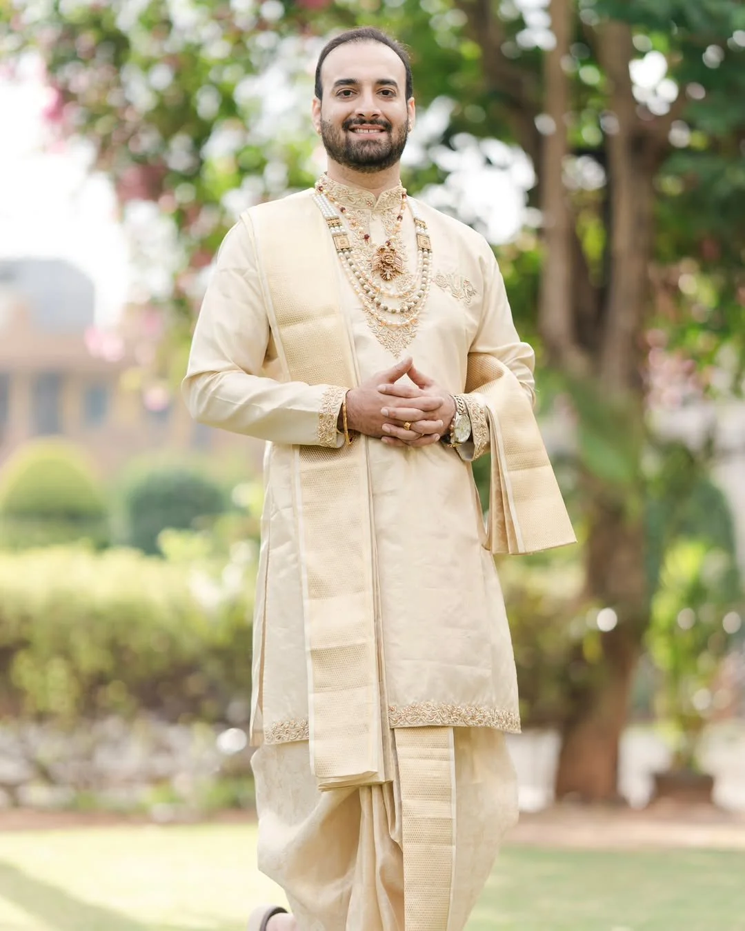 A man dressed in traditional Indian cream-colored wedding attire with gold embroidery, wearing multiple gold and pearl necklaces, rings, bracelet, and watch, standing outdoors in a garden with trees and blurred greenery in the background.