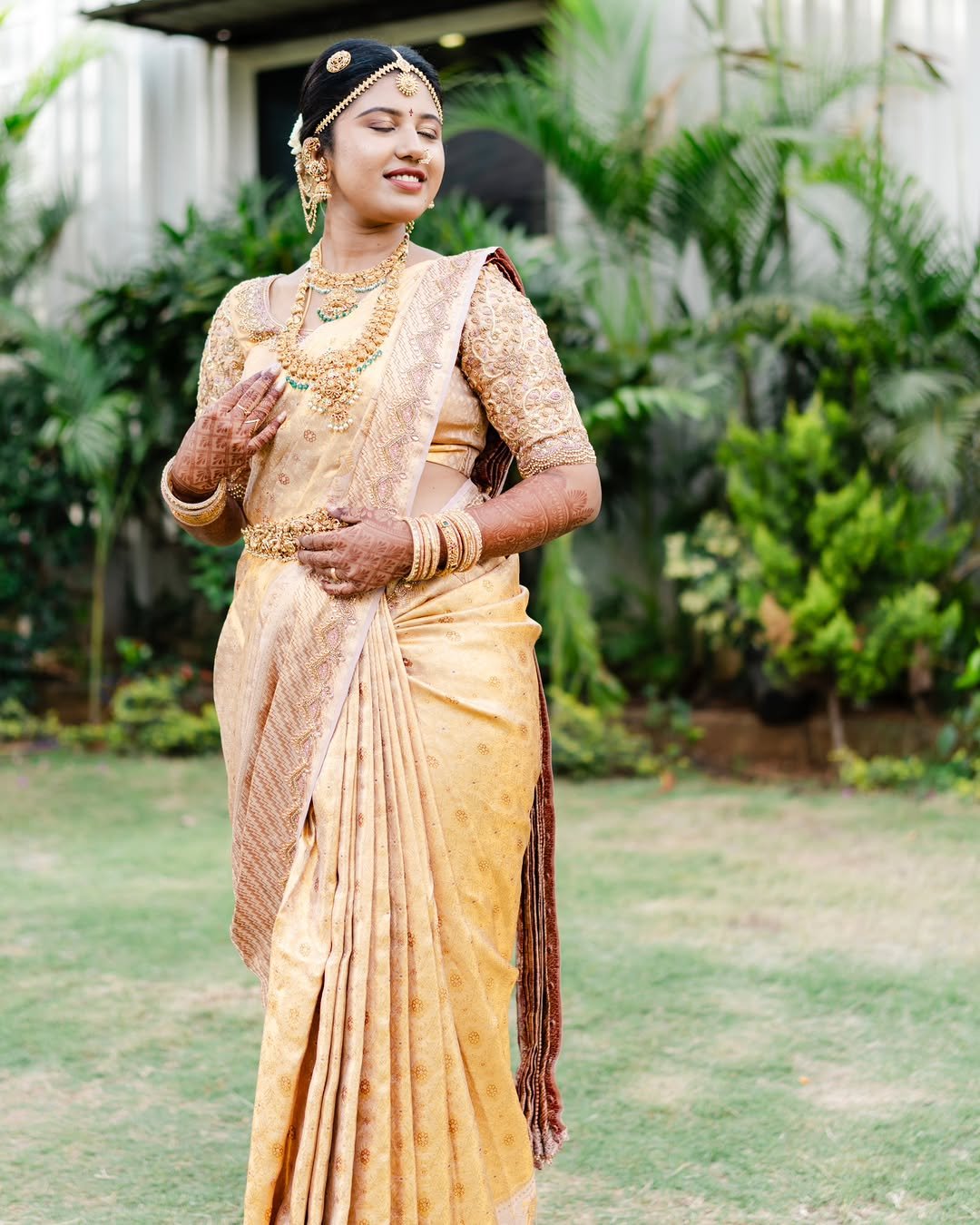 A woman dressed in traditional Indian attire, wearing gold jewelry, a saree, and bangles, standing outdoors with greenery in the background.