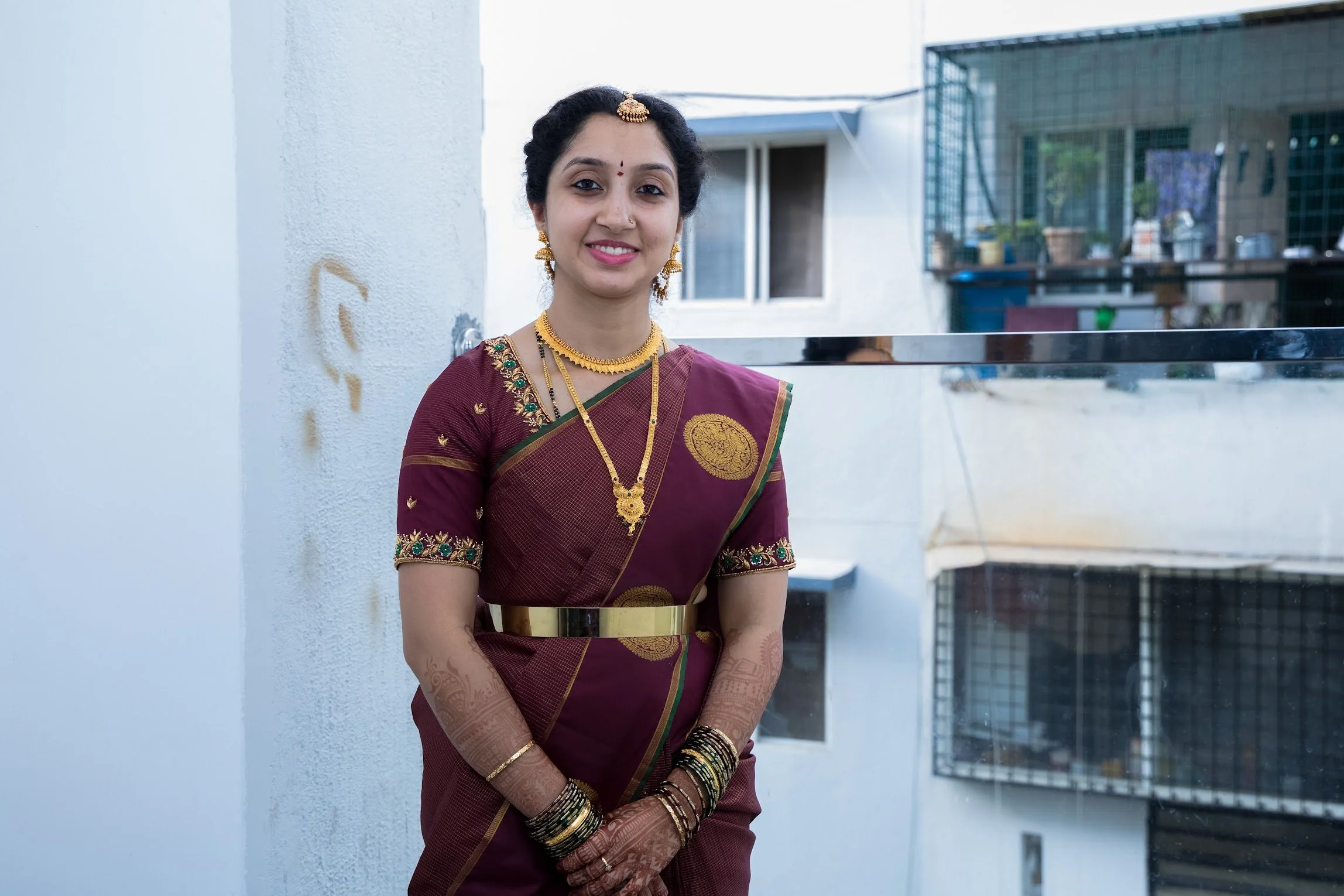 A woman in traditional Indian attire, with jewelry and henna on her hands, standing outdoors in front of a white wall and balcony.