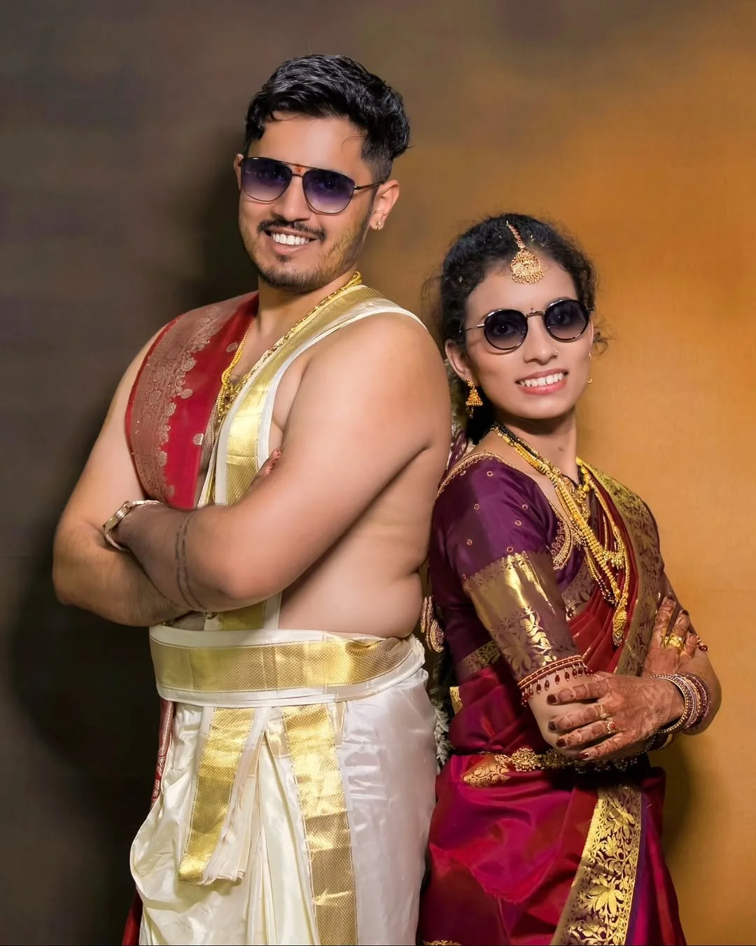 A smiling man and woman wearing traditional Indian clothing and sunglasses, standing back to back with arms crossed, against a yellow and brown background.