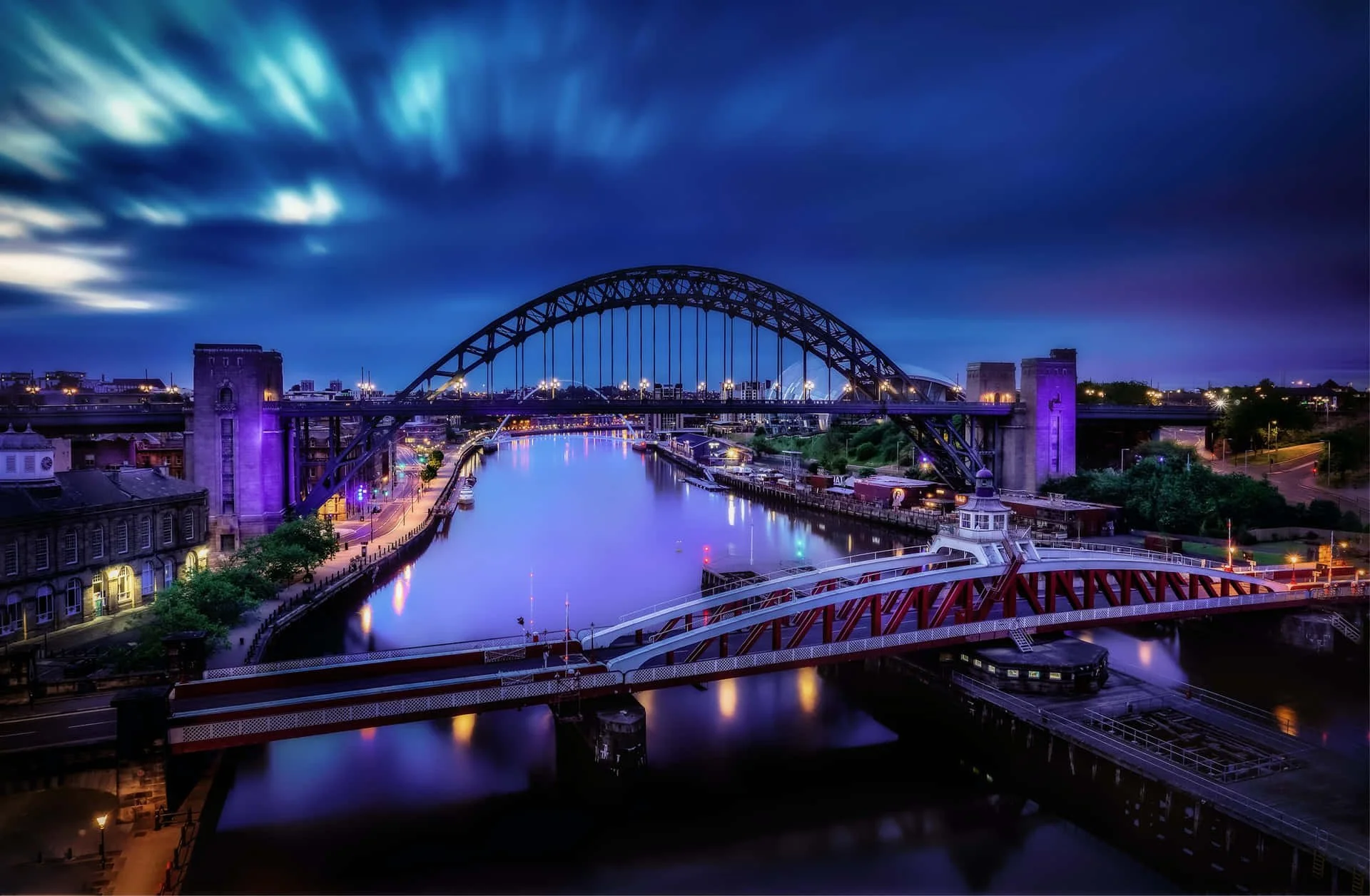 Night view of the Tyne Bridge over the River Tyne in Newcastle upon Tyne, with illuminated buildings and city lights reflecting on the water, under a dark cloudy sky.