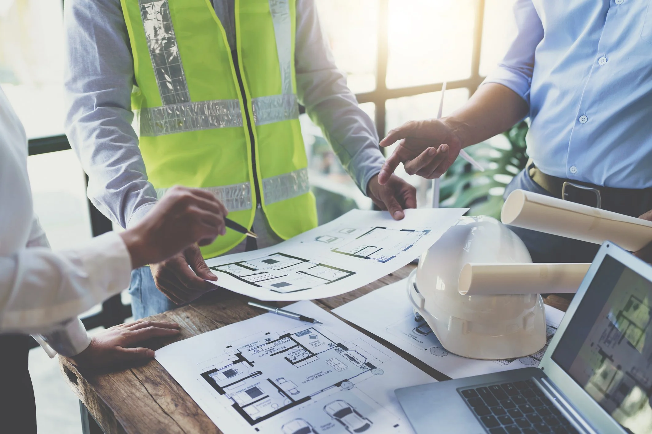 People reviewing architectural plans and blueprints on a wooden table, with a person wearing a high-visibility safety vest.