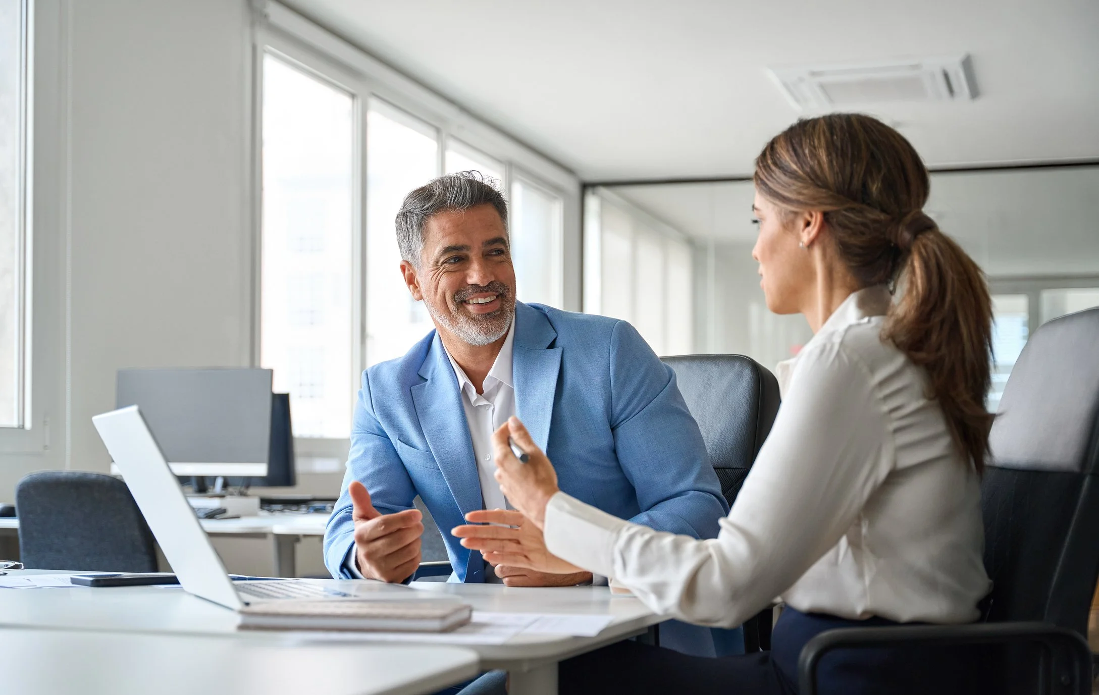 A man in a light blue suit and a woman in a light-colored blazer are engaged in conversation in a modern office with large windows. The man has gray hair and a beard, smiling, while the woman has brown hair tied back, holding a pen. They are sitting at a table with laptops and papers.