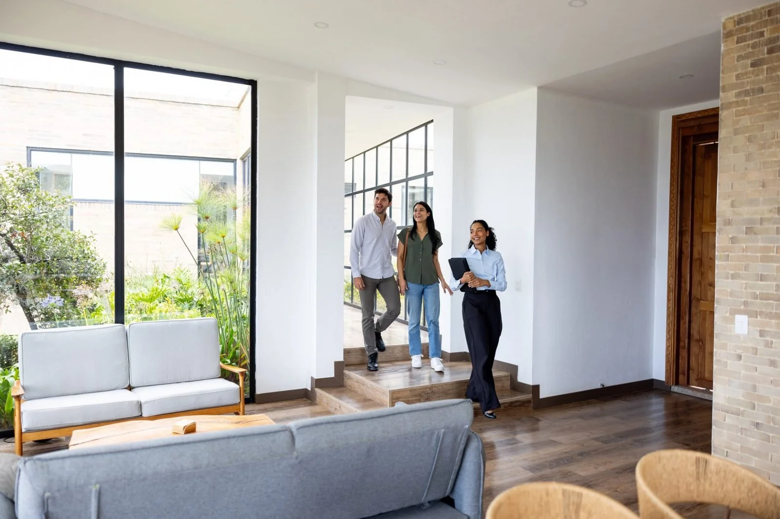 Three people, a man and two women, standing in a modern, well-lit living room with large windows and greenery outside, talking to a real estate agent.