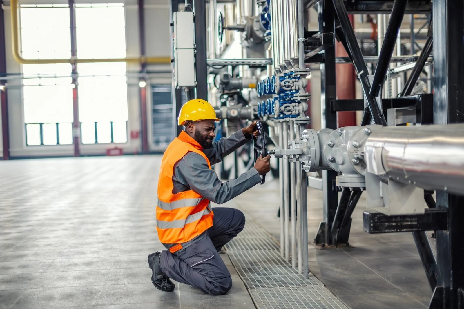 A worker wearing a yellow hard hat and orange safety vest kneels while inspecting or working on large industrial pipes in a factory or industrial setting.