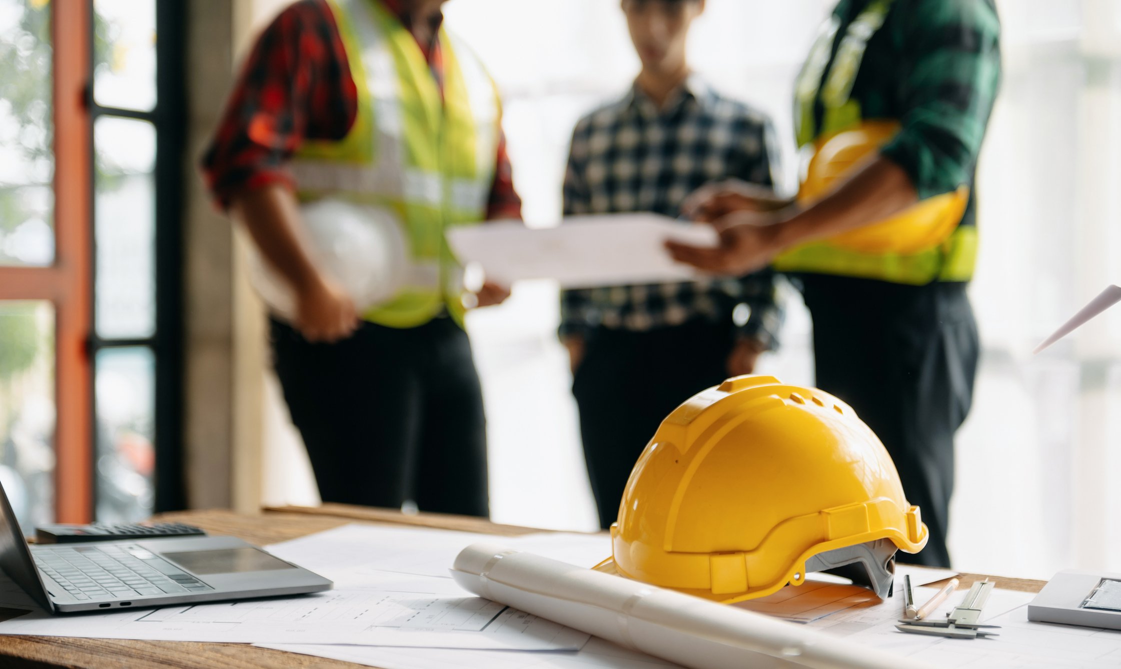 Construction helmets and blueprints on a table with people in safety vests discussing in the background.