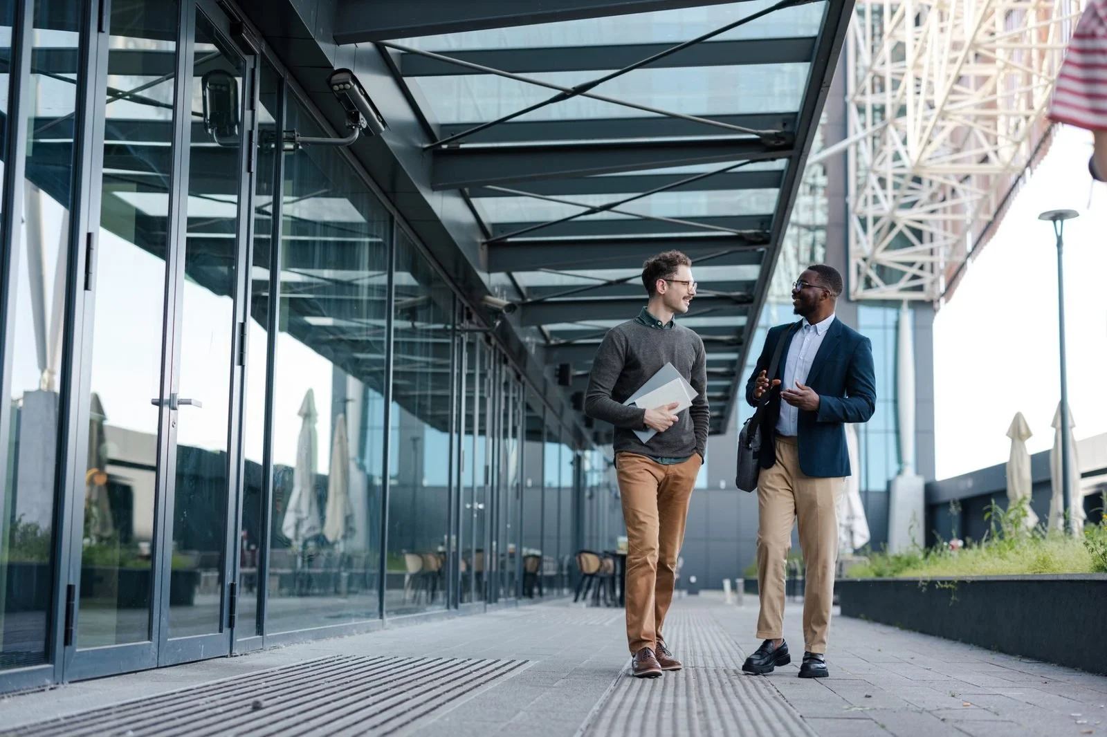 Two men in business attire walking and talking outside a modern glass office building on a clear day.