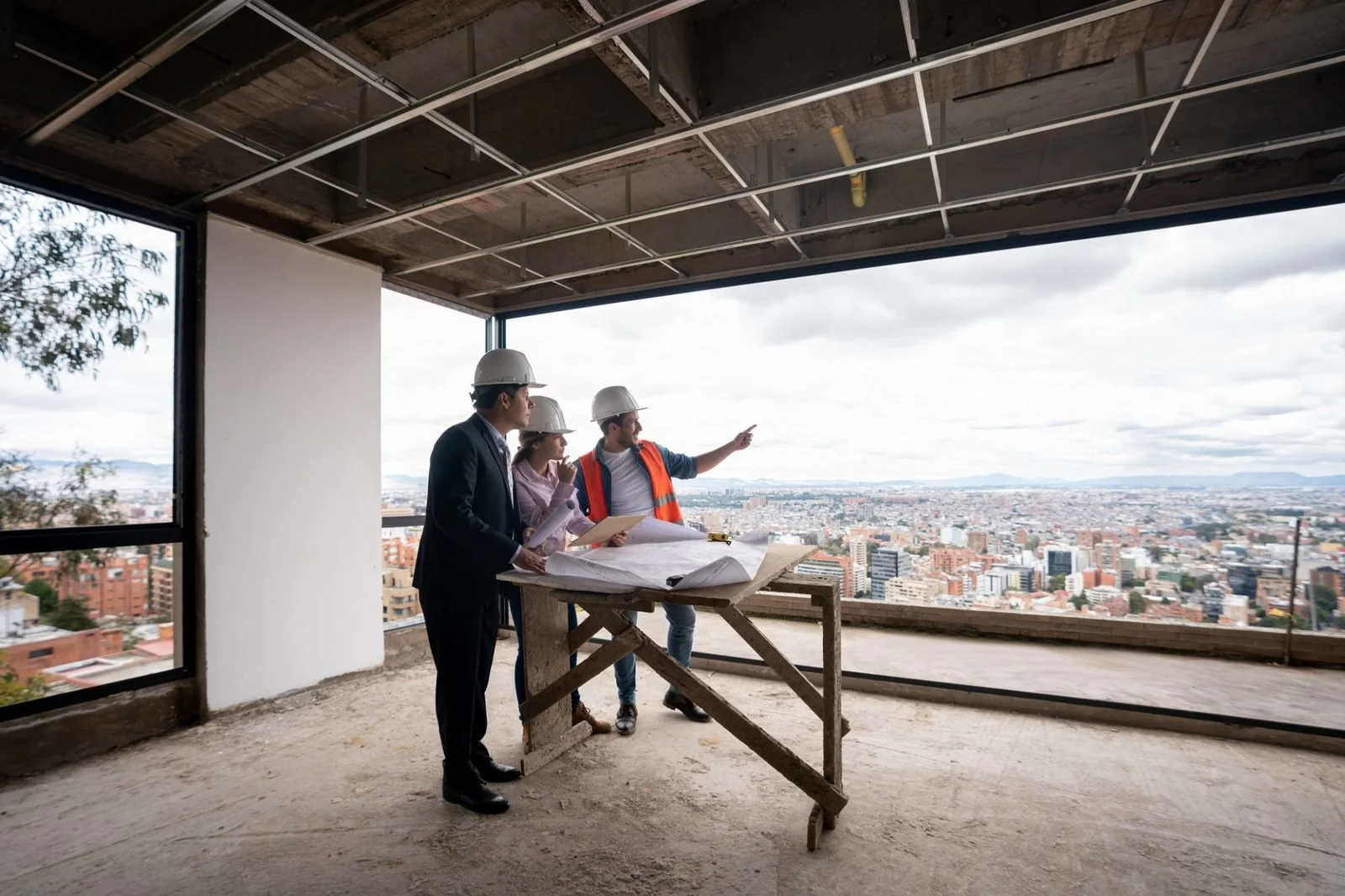 Three construction professionals, two men and one woman, wearing safety helmets and a safety vest, reviewing building plans on a construction site with a cityscape view in the background.
