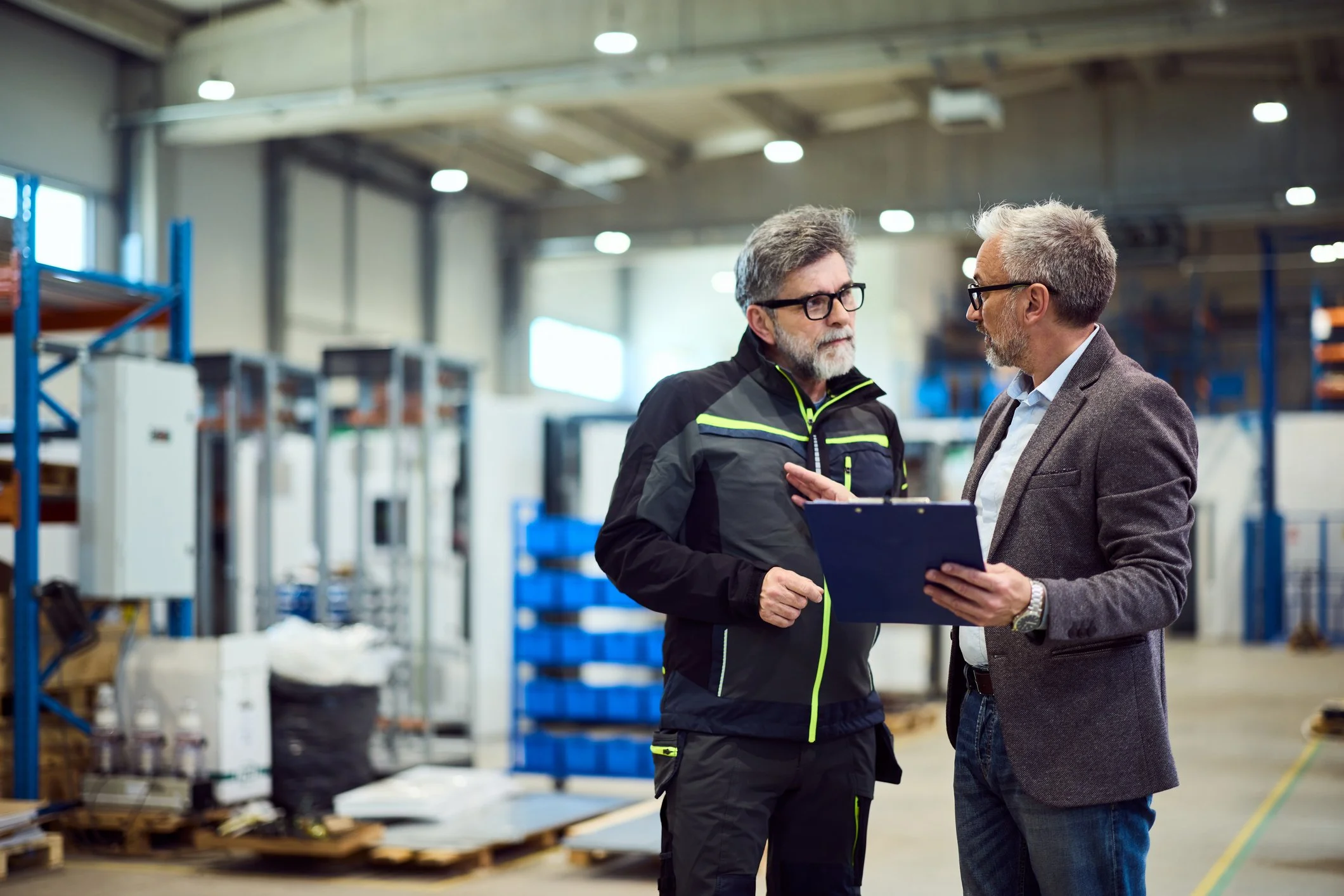 Two men having a discussion in an industrial warehouse. One is wearing a black jacket and glasses, holding a clipboard, and the other is wearing a brown blazer with a white shirt and glasses. The warehouse has metal shelves, boxes, and welding equipment in the background.