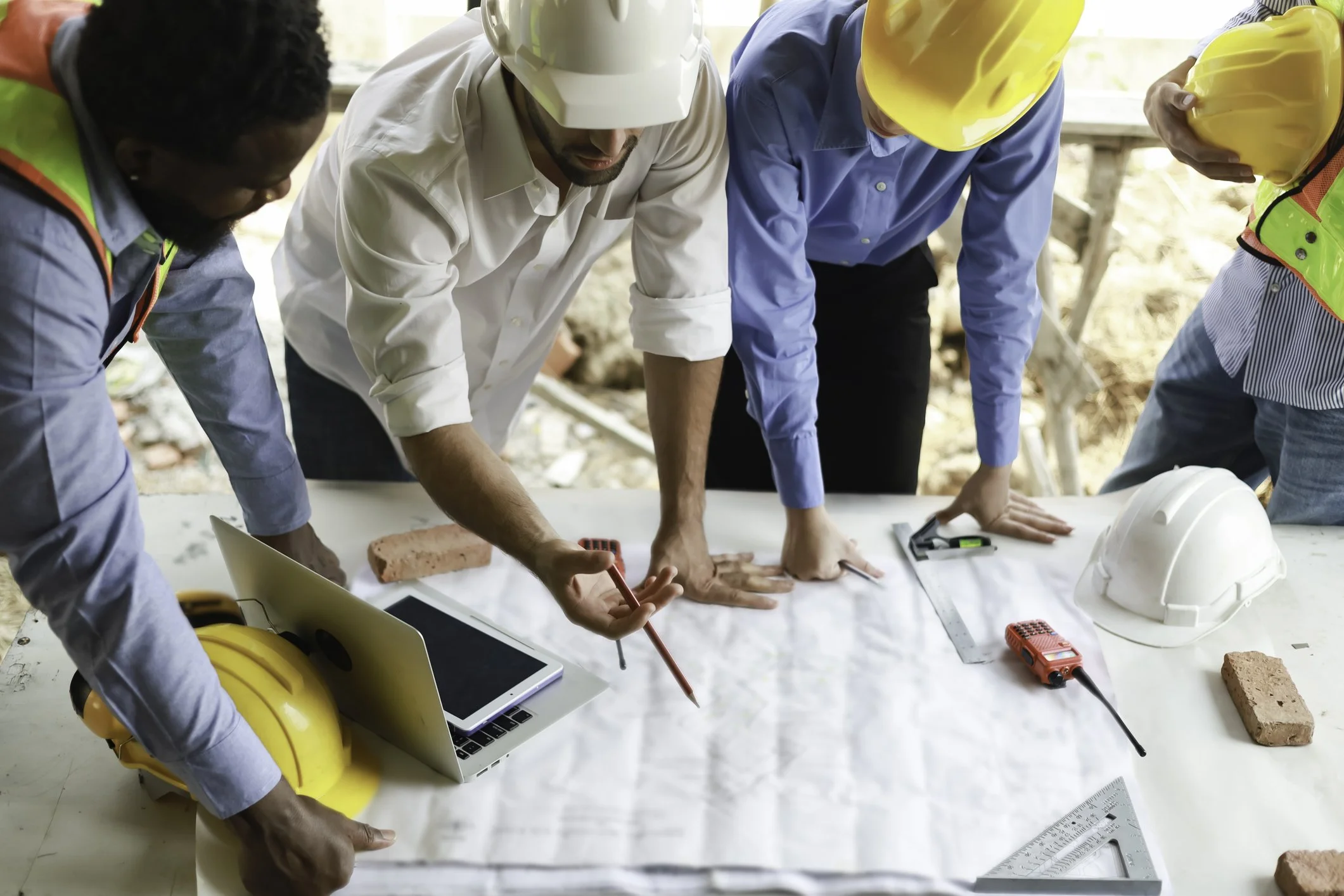 Group of construction professionals, some wearing safety helmets, examining plans and working on a construction site.