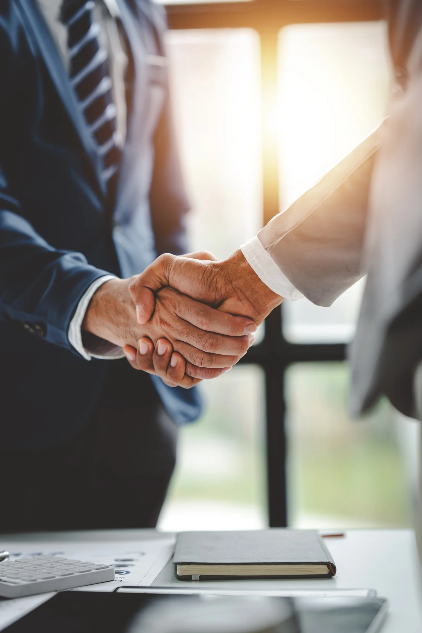 Two people shaking hands over a desk with a closed notebook, calculator, and papers, signifying a business agreement or greeting.