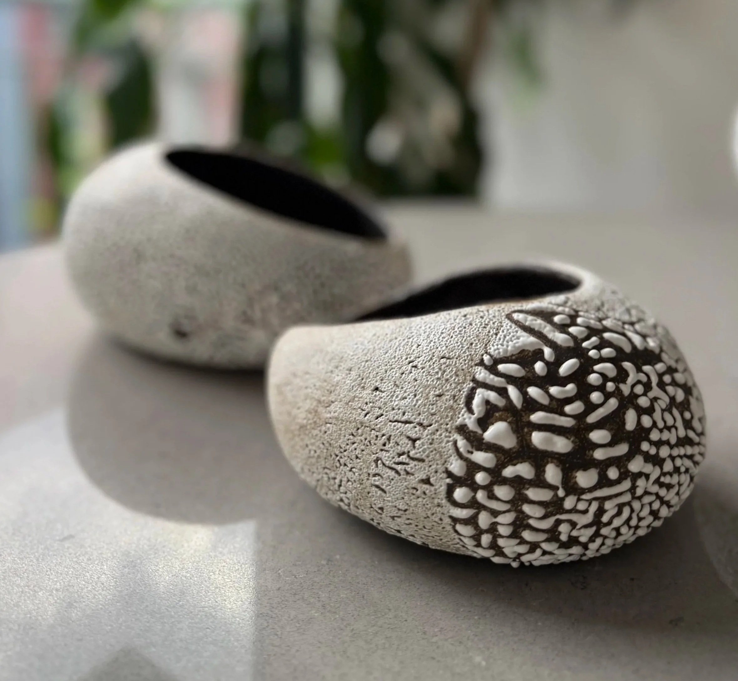 Close-up of a round, textured black and white patterned object, possibly a piece of art or decor, with a blurred background featuring green and light-colored elements.