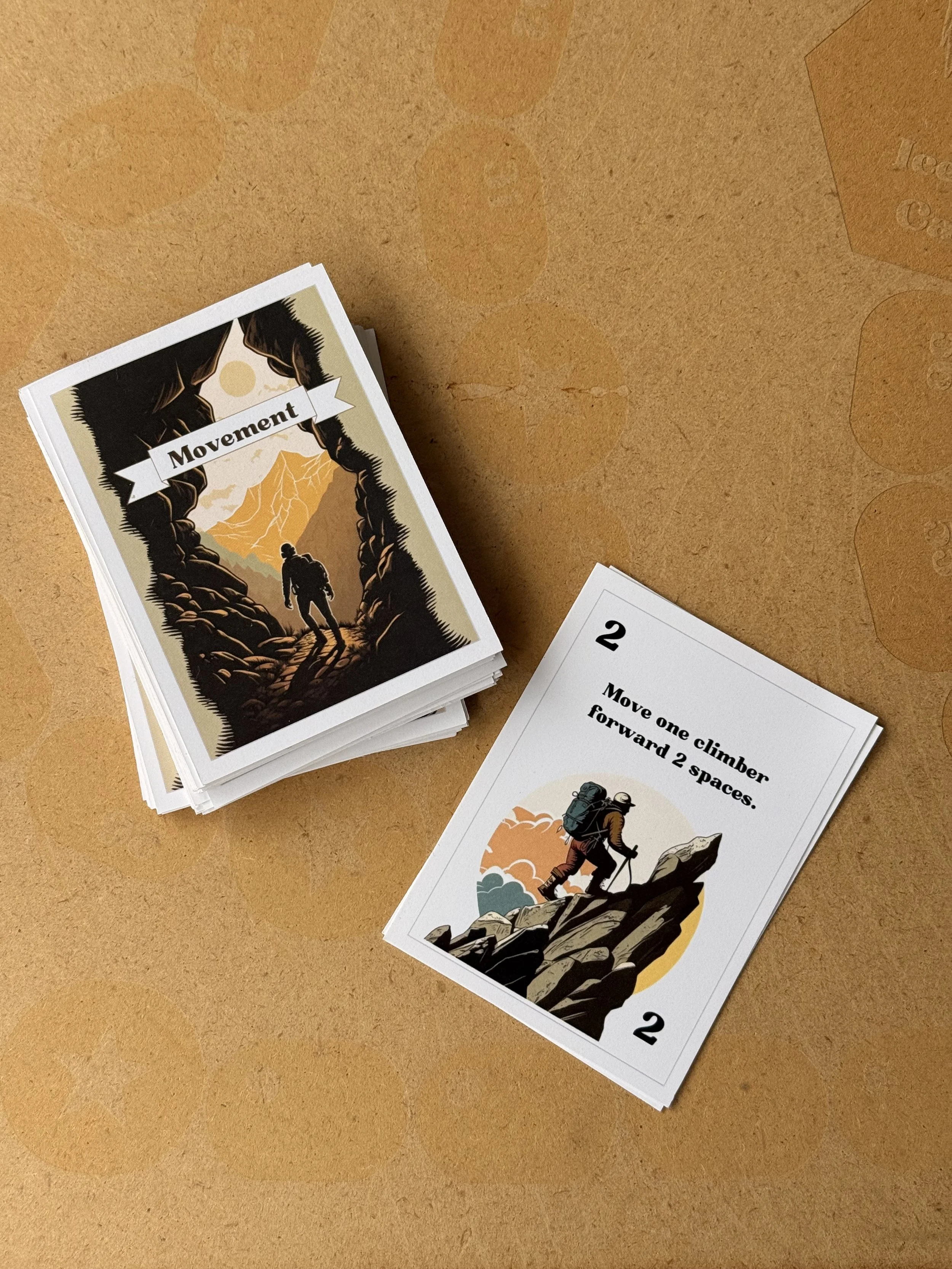 A stack of illustrated cards with outdoor adventure themes on a brown surface. The top card shows a hiker climbing a mountain with a sun or moon in the background, and the word 'Movement.' Another card visible features a hiker stepping over rocks wit