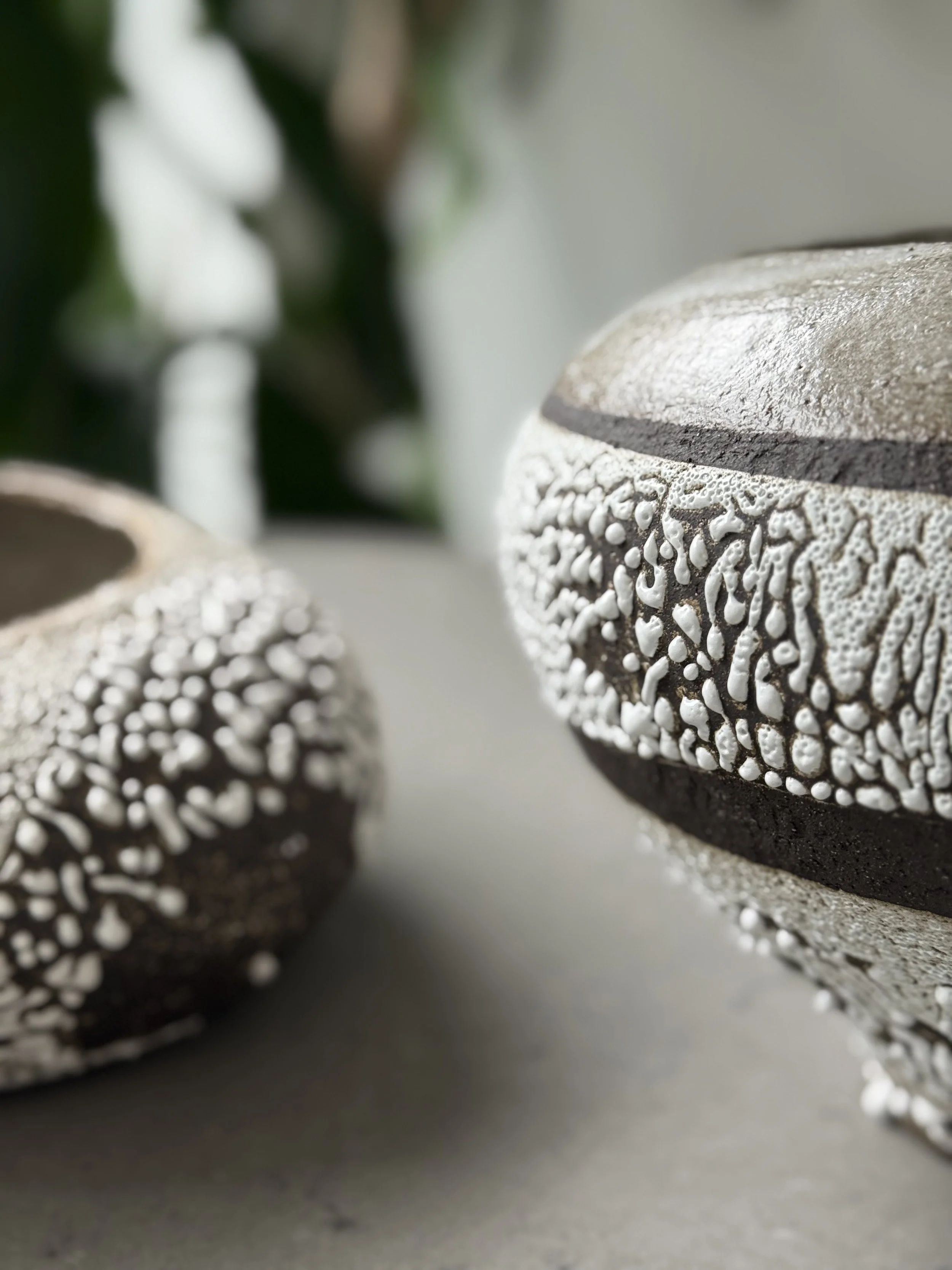 Close-up view of two decorative vases with textured surfaces, one in the foreground and one in the background, with blurred greenery behind them.