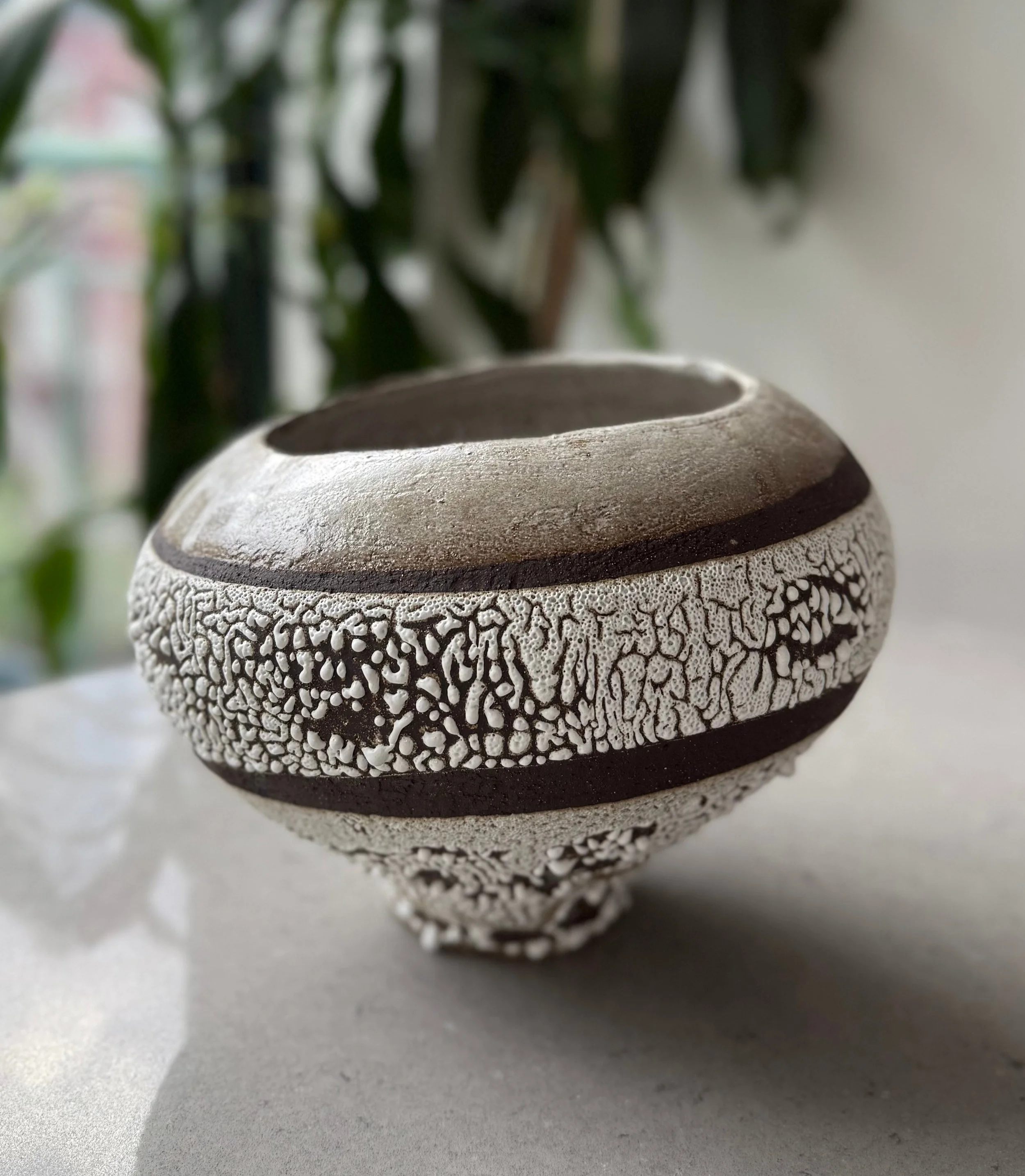 Close-up of a decorative ceramic vase with intricate textured patterns, placed on a flat surface with a blurred background of green leaves.