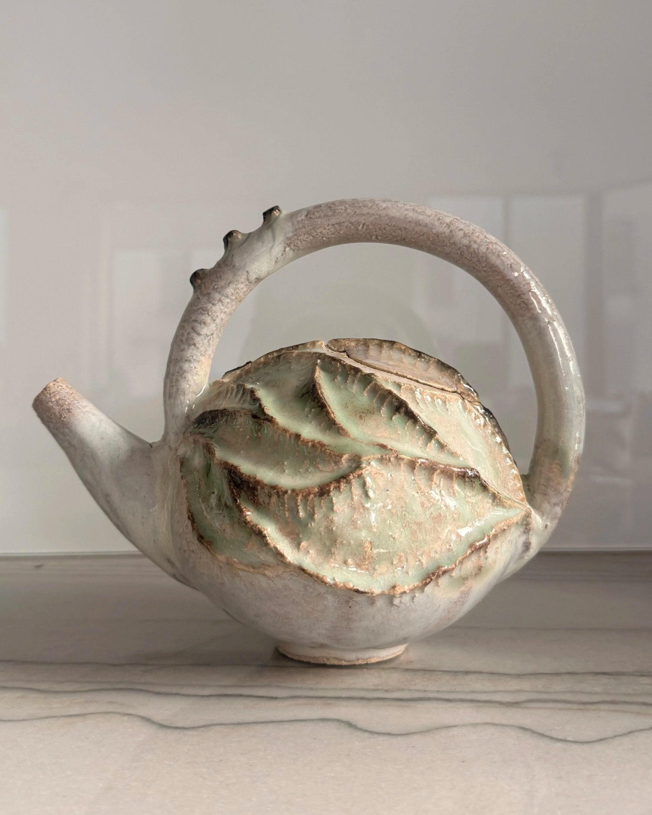 A decorative ceramic teapot with a white, pink, and green glaze, designed with a leaf motif on its body and a curved handle, placed on a light wooden surface.