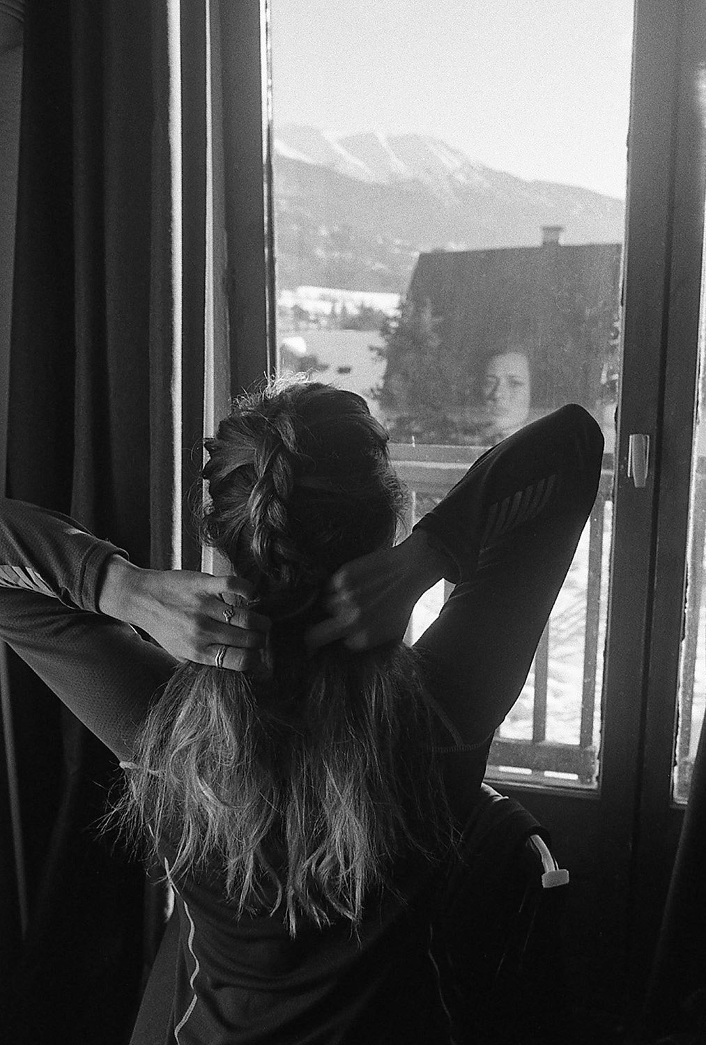 A woman with braided hair seen from behind, sitting by a window, tying her hair back.