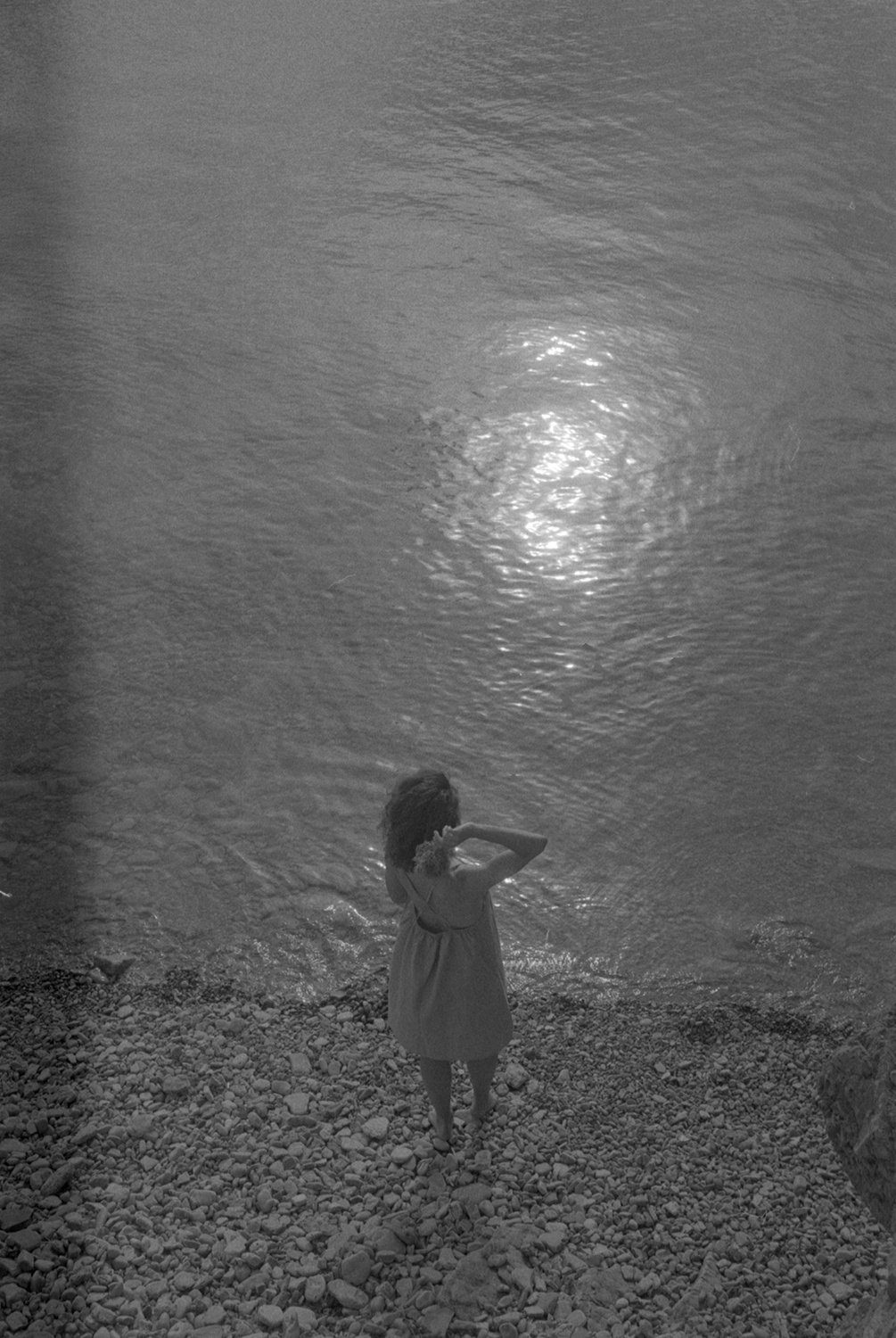 A woman in a dress standing on a rocky shoreline, looking out at the water with the reflection of the moon or sun on the surface.