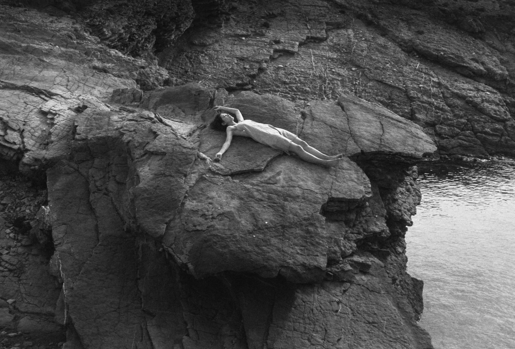 A woman lying on a large rock formation near water, surrounded by rocky cliffs.