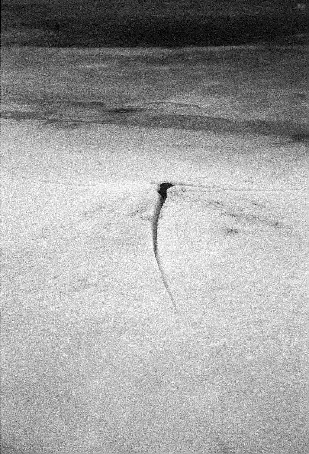 A snow-covered landscape with a single dark crevasse or crack in the snow, leading towards a dark opening or tunnel, under a cloudy sky.