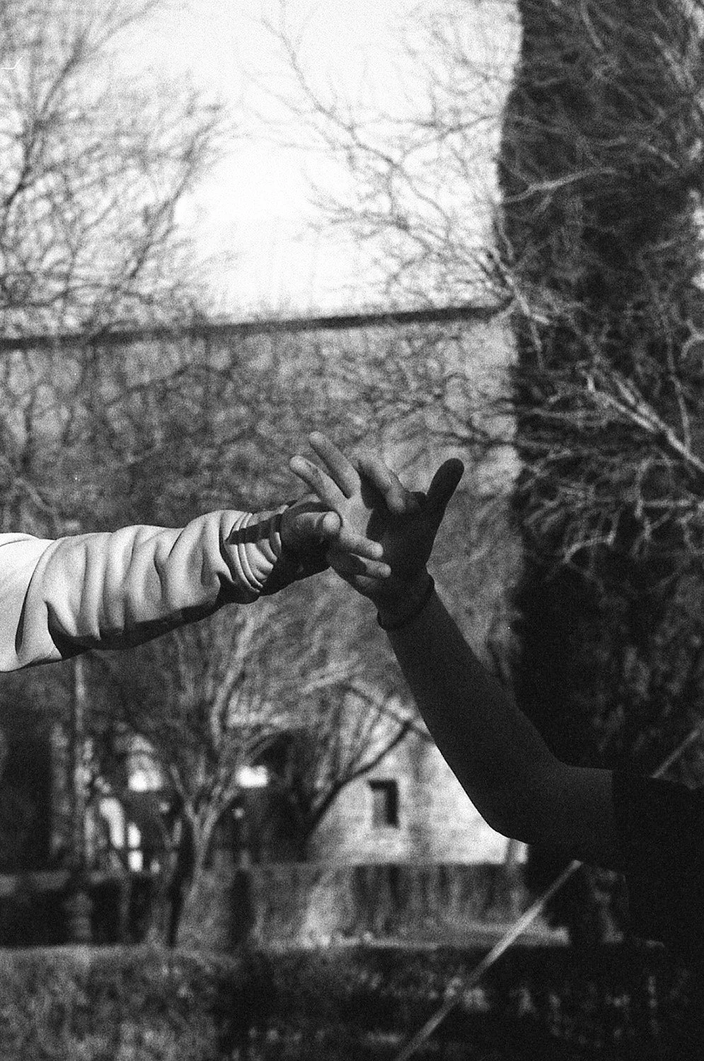 Two hands, one in a glove, reaching towards each other, against a backdrop of bare trees and a building in a black and white photo.