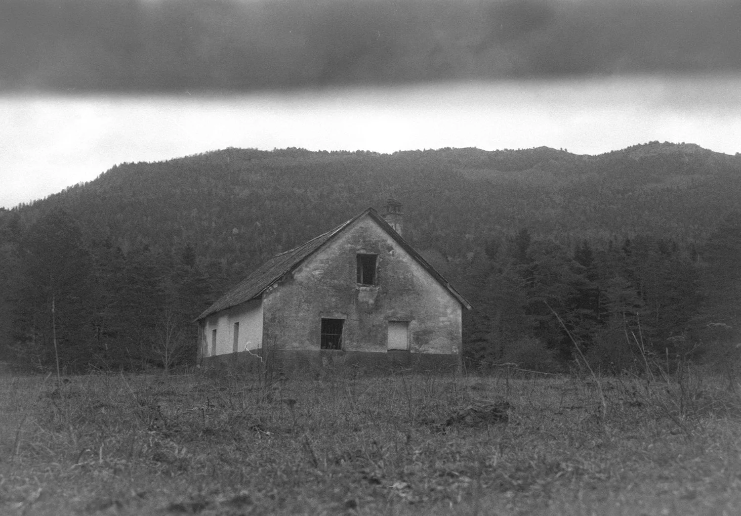 Black and white photo of an abandoned house with broken windows, surrounded by tall grass and trees, with a mountain range and overcast sky in the background.