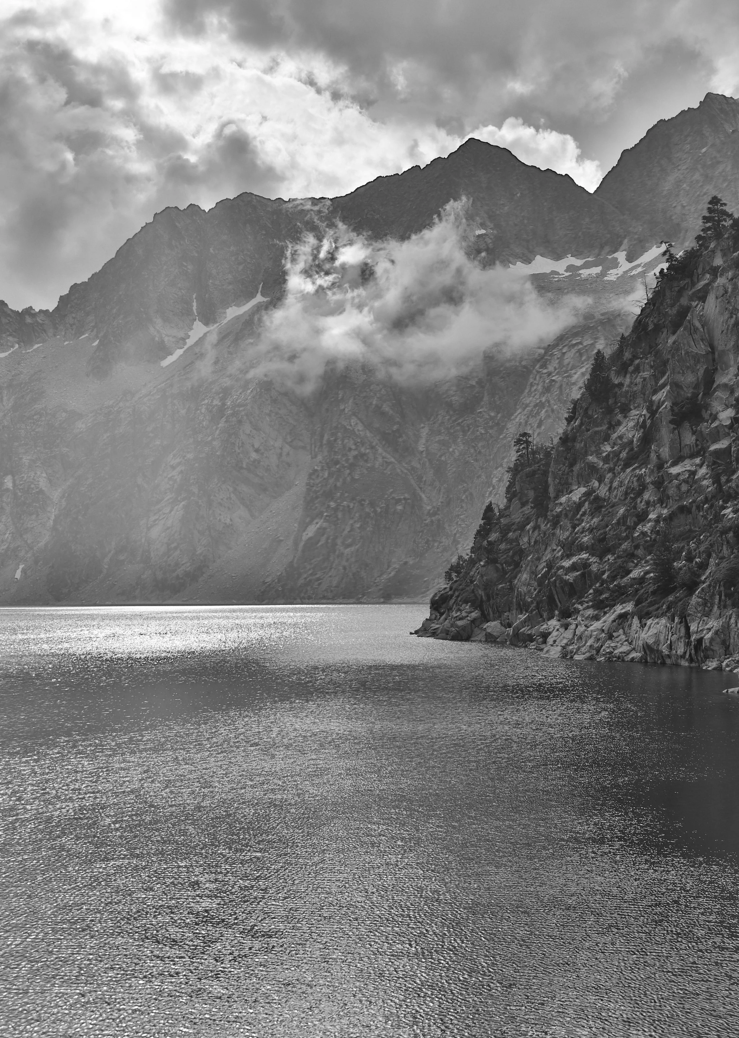 Black and white photo of a mountain landscape with water in the foreground, rocky slopes on the right, and clouds above the peaks.
