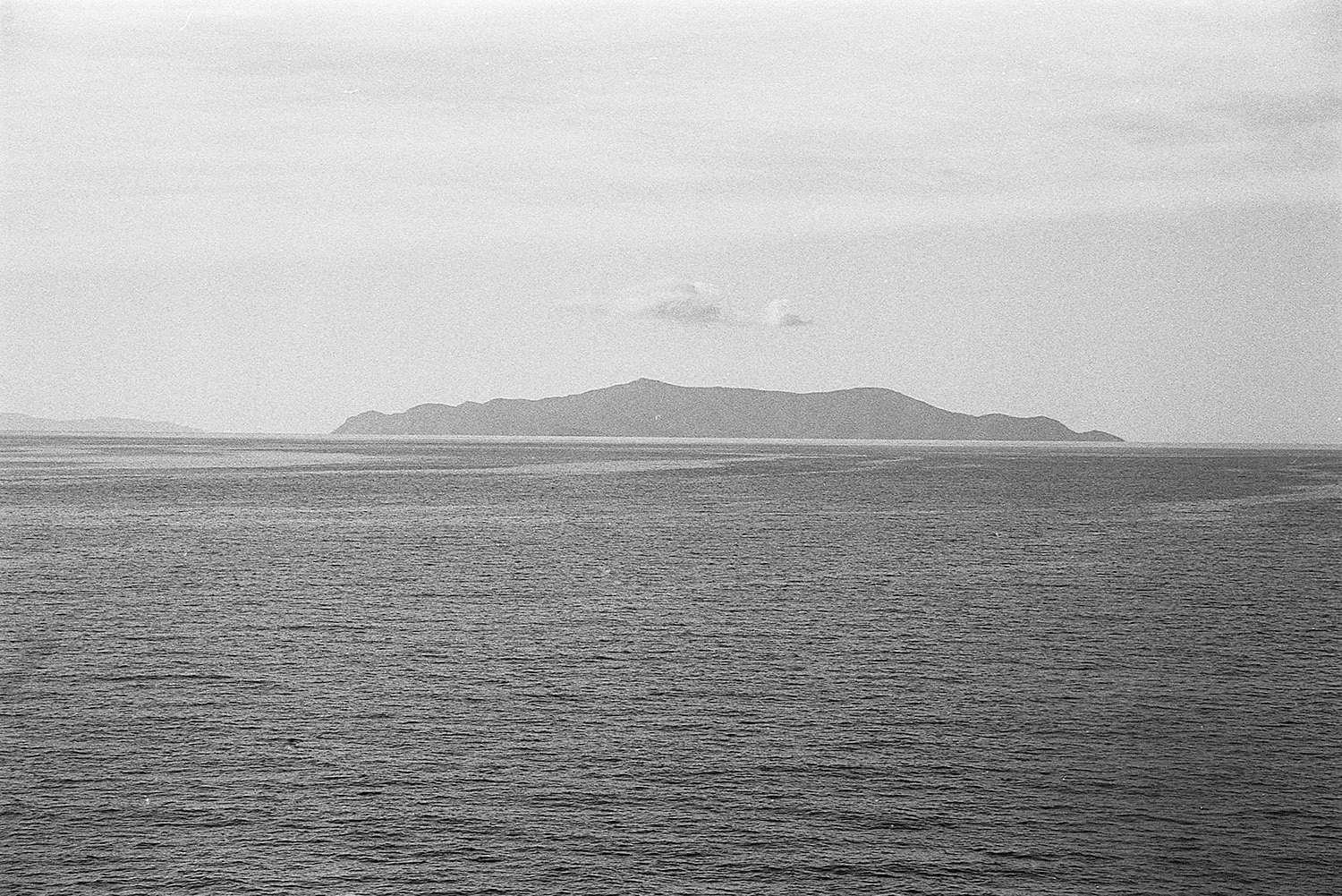 Black and white photo of a body of water with a mountainous island in the background under a partly cloudy sky.