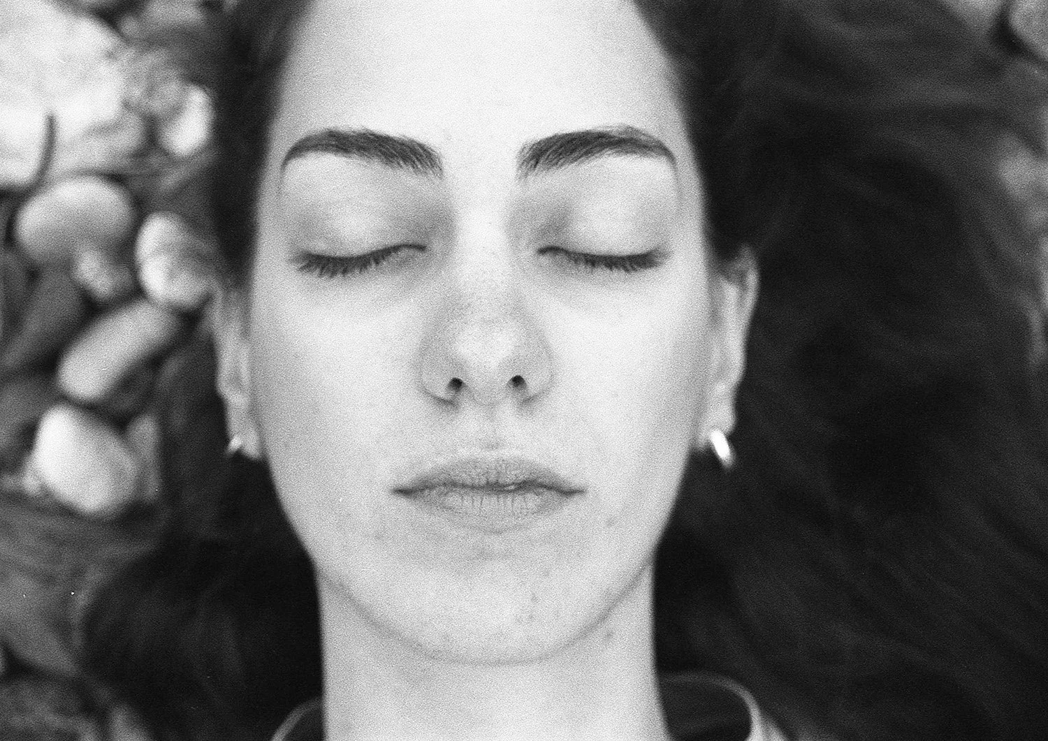 A woman with closed eyes, lying down with dark hair spread around her, touching her face with one hand.