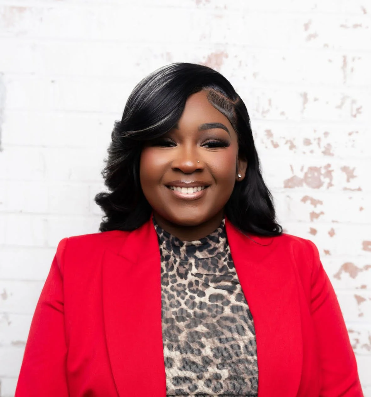 Portrait of a young Black woman with black hair, wearing a red blazer and a leopard print top, smiling in front of a white brick wall.
